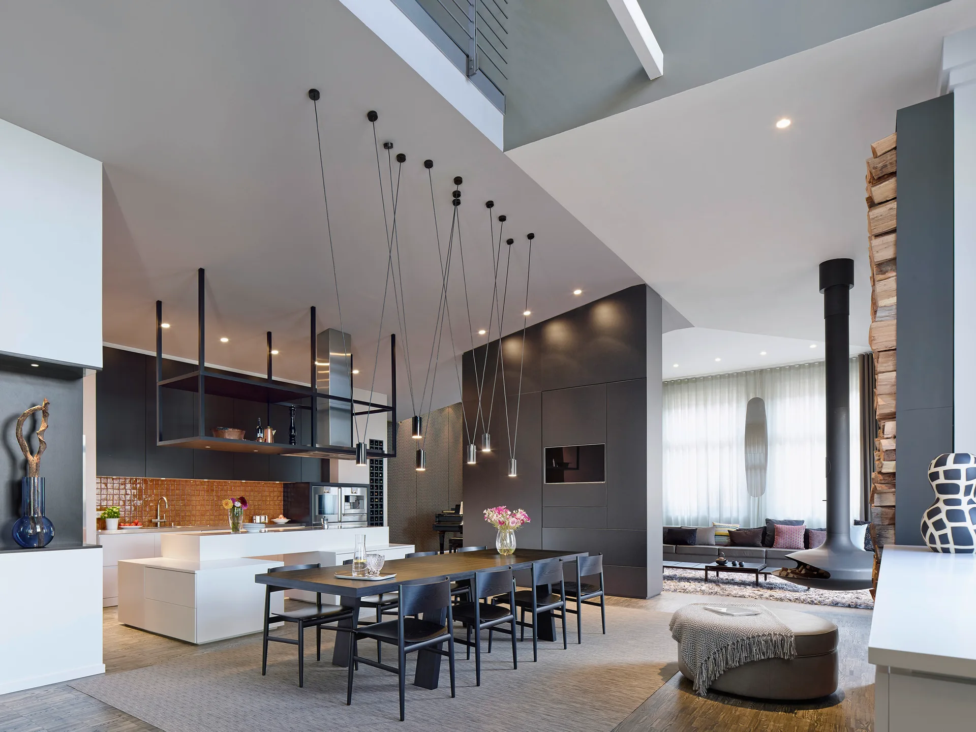 A modern open-plan dining space features a black table and chairs, slim hanging pendant lights, a white kitchen island, and a suspended black fireplace.