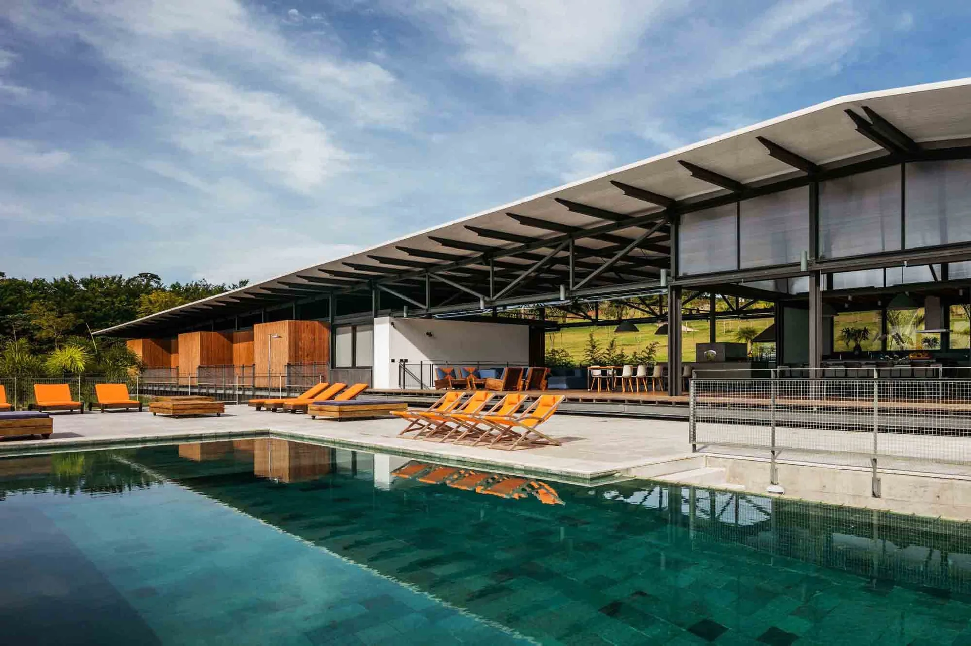A rectangular turquoise swimming pool sits next to a modern open pavilion with orange lounge chairs and an exposed steel roof structure.