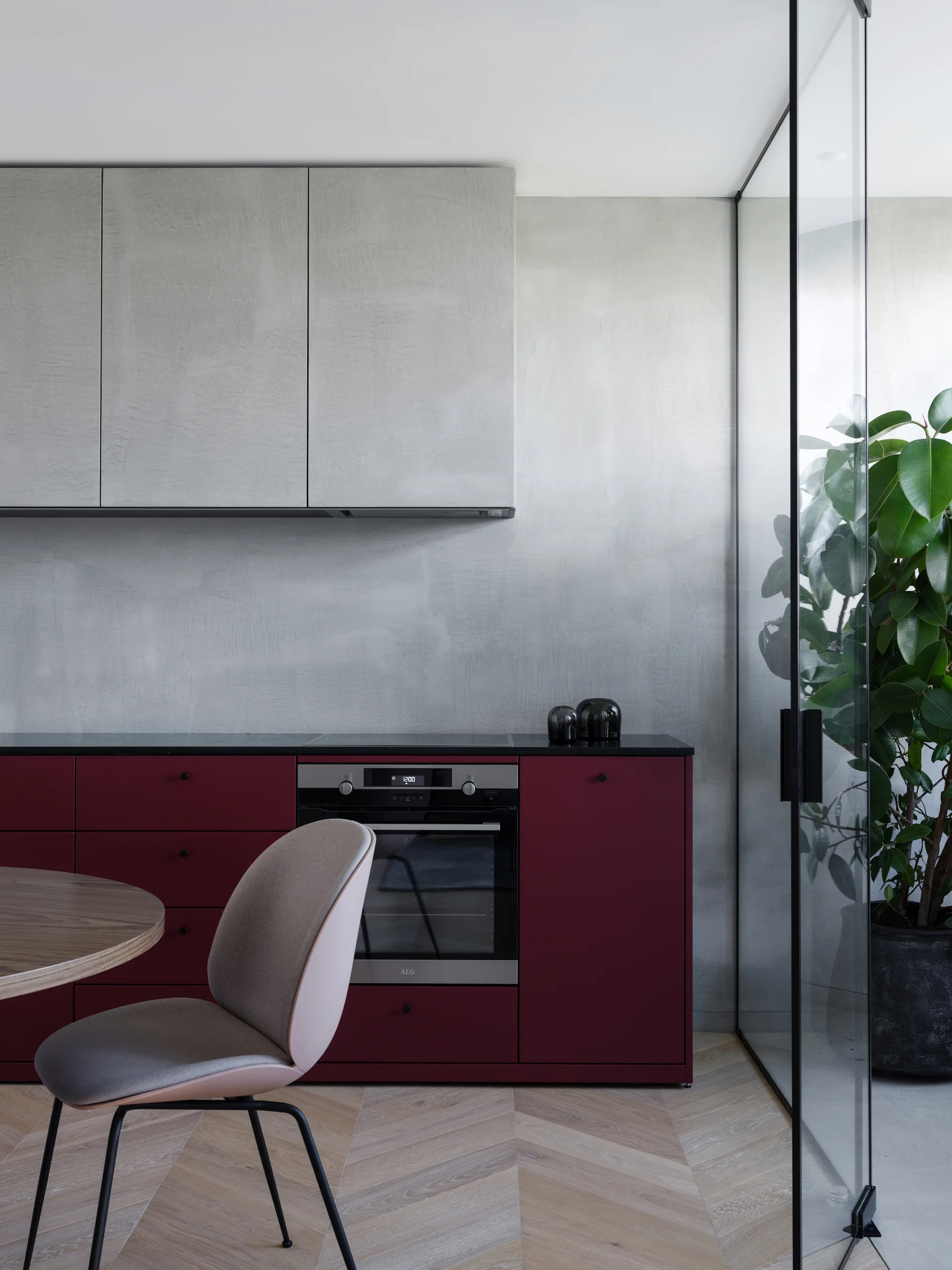 A modern kitchen features matte burgundy lower cabinets, textured gray upper cabinets, herringbone wood floors, and a black-framed glass wall.