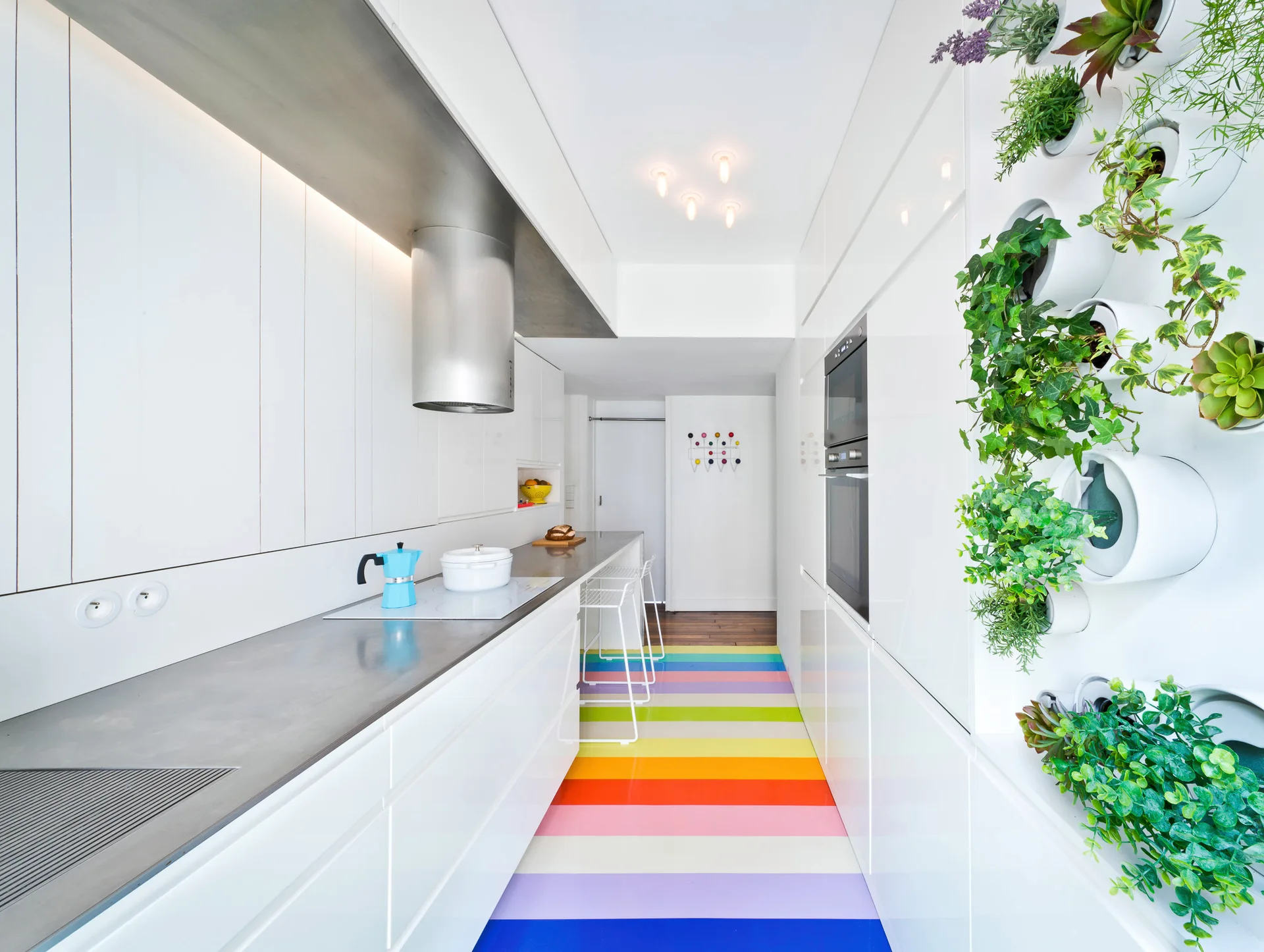 A narrow white galley kitchen features a colorful rainbow-striped floor, sleek white handleless cabinets, and a wall-mounted vertical garden.