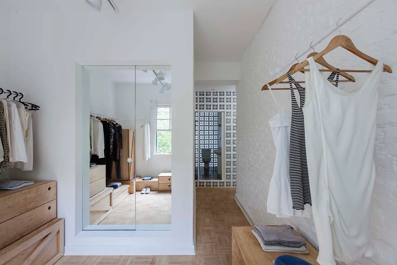 A bright dressing room featuring white painted brick walls, natural wood drawers, and a large mirrored partition reflecting a window.