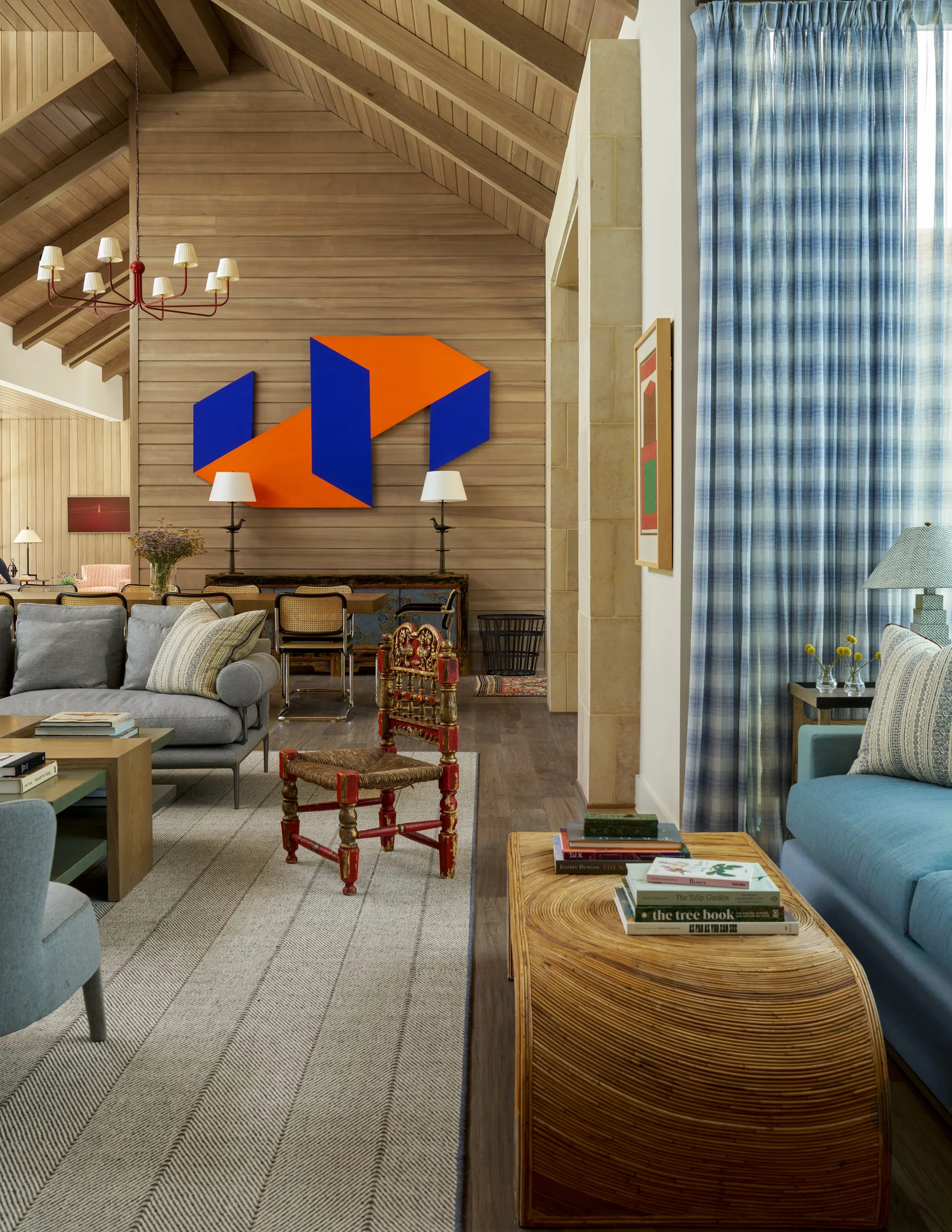 An eclectic living room featuring a vaulted wood-paneled ceiling, a grey sofa, blue plaid curtains, and a large blue and orange geometric art piece.