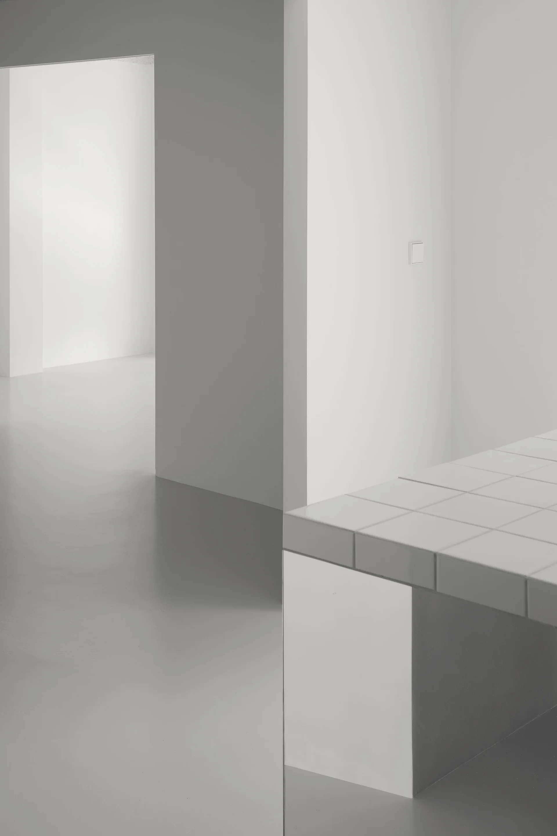 A minimalist interior featuring plain white walls, a smooth gray floor, and the corner of a white tiled table in the foreground.