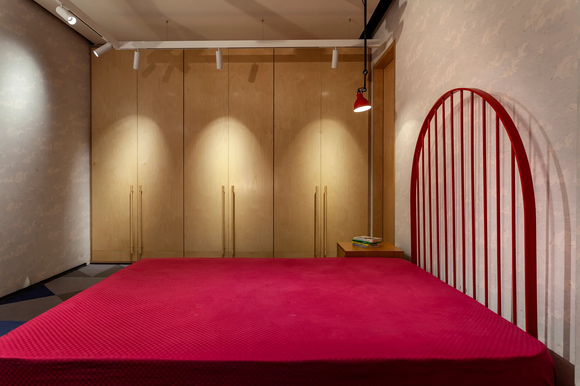 A modern bedroom featuring a bed with a vibrant red arched headboard and a full wall of light-toned wood cabinetry illuminated by overhead track lights.