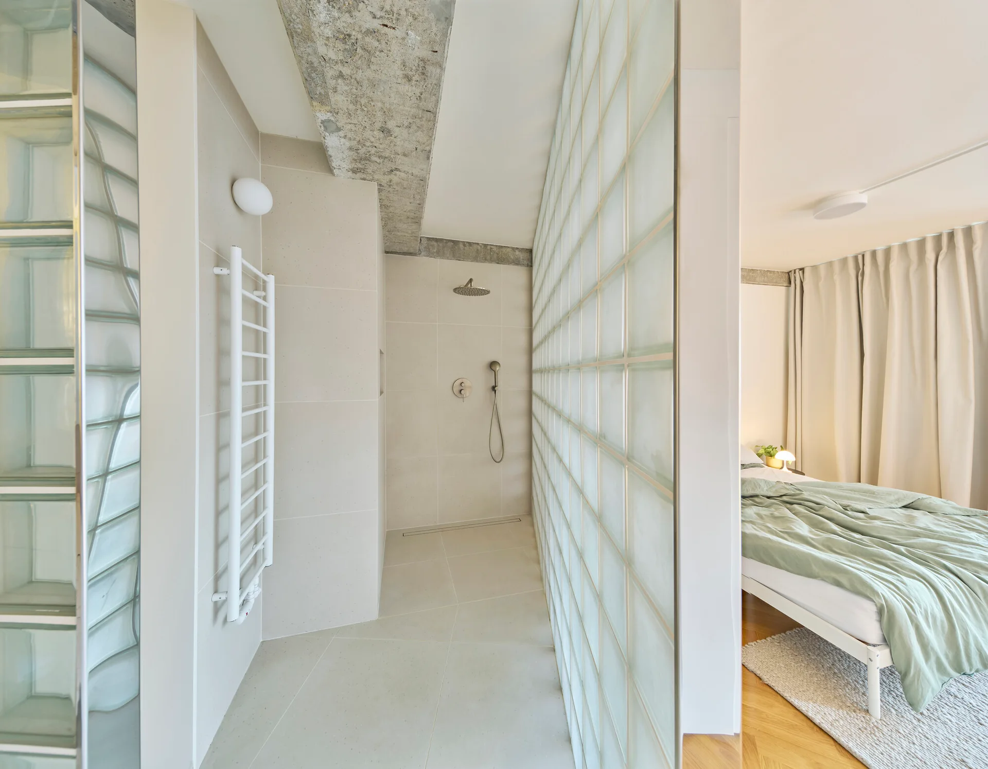 A glass block wall divides a walk-in tiled shower with a white towel radiator from a modern bedroom featuring green bedding.
