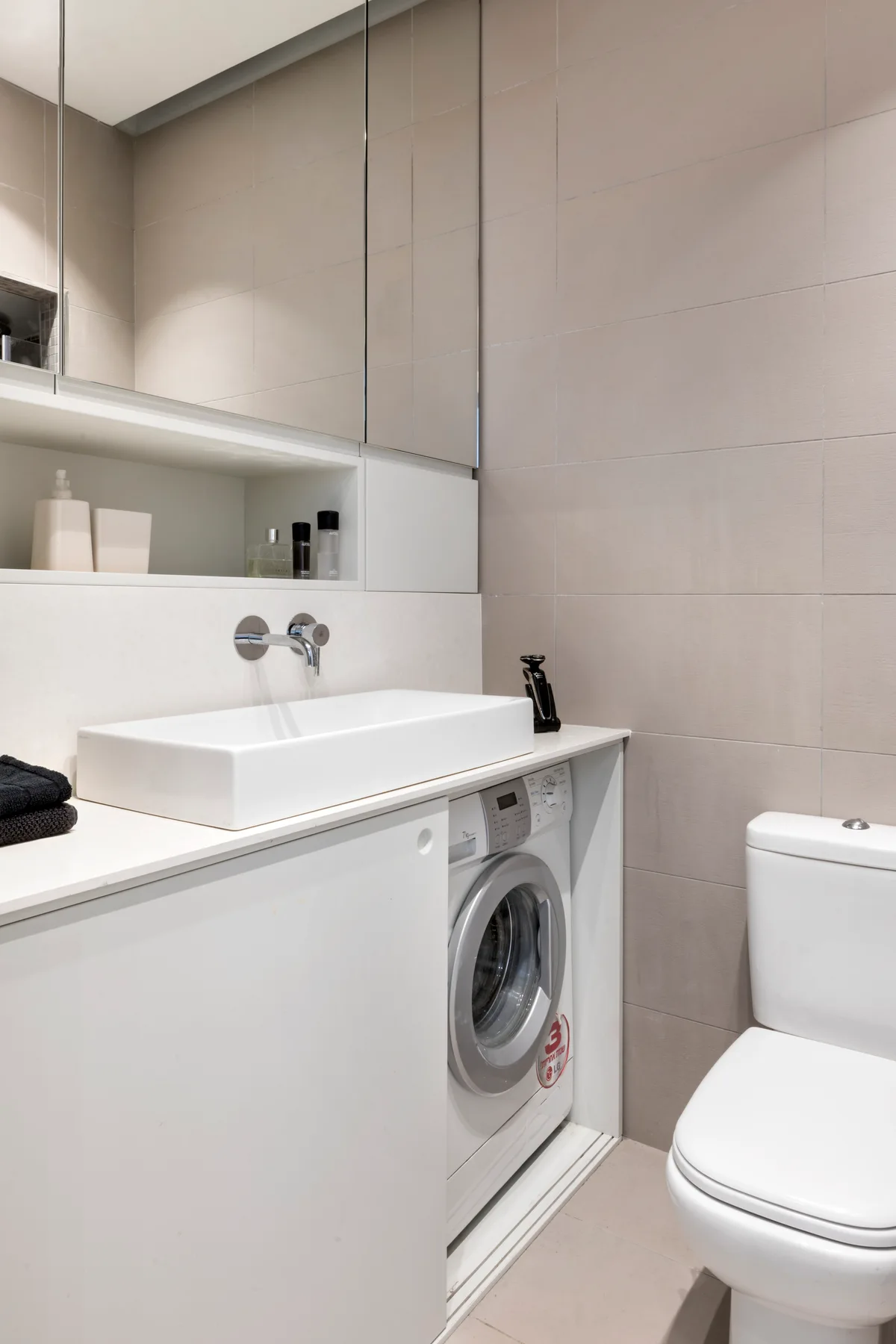 A modern bathroom features a white vanity housing an integrated washing machine, a rectangular vessel sink, a wall-mounted faucet, and a toilet.