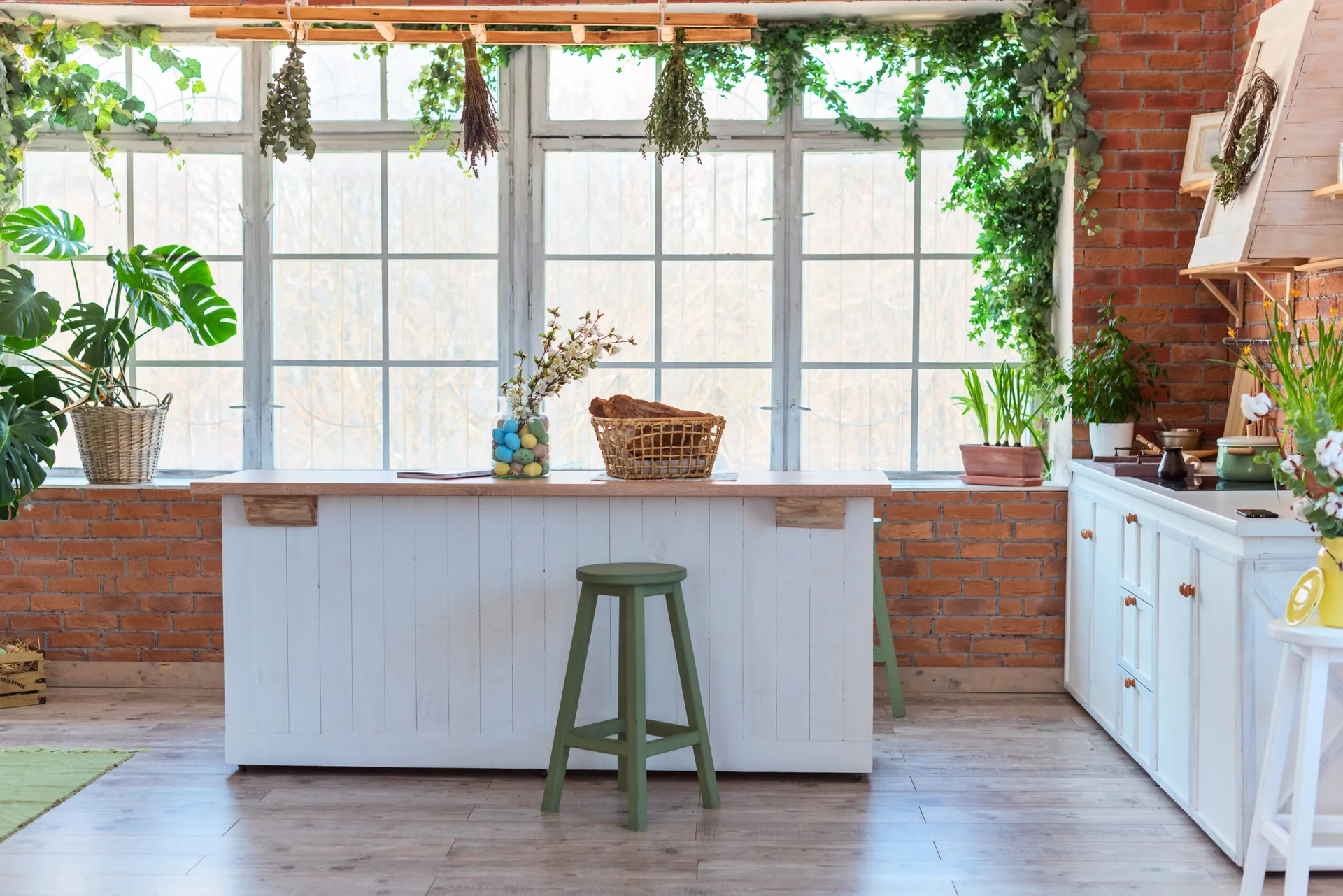 A rustic kitchen features a white wood island with a green stool, set against an exposed brick wall and large windows draped in leafy green vines.