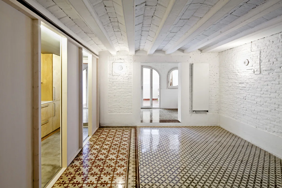 An empty room features white painted brick walls, a vaulted ceiling with exposed beams, and two distinct patterns of geometric ceramic floor tiles.