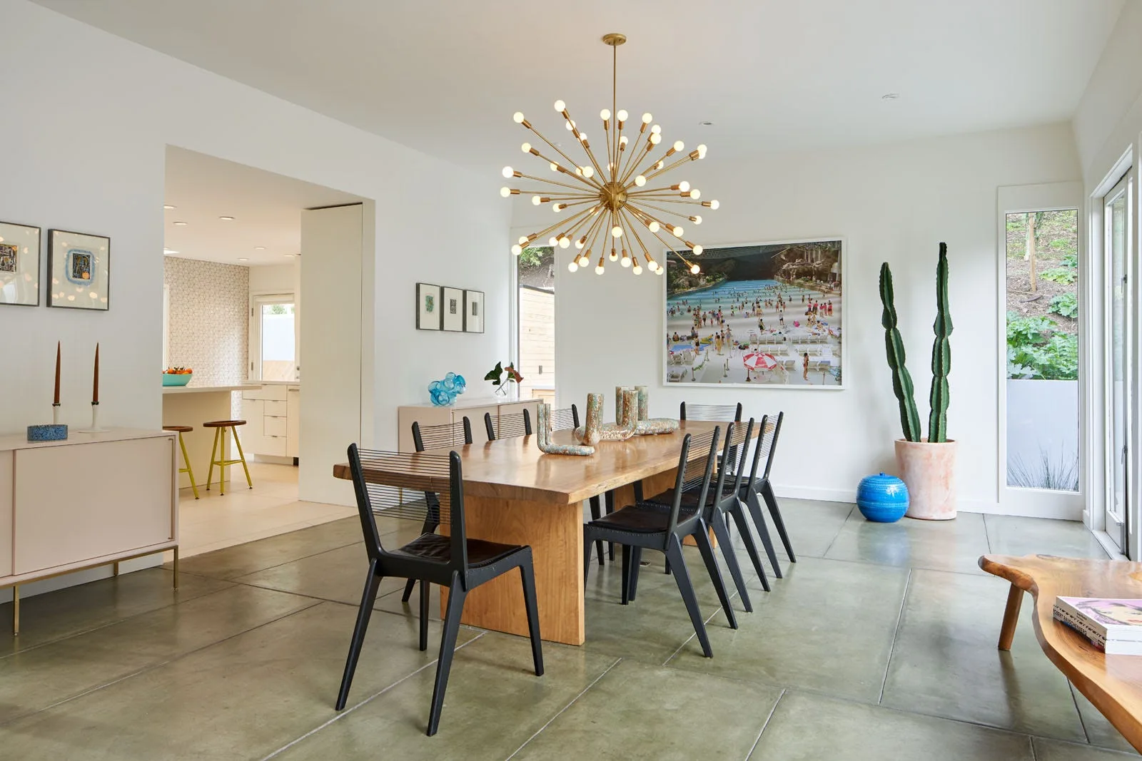 A modern dining room featuring a long wood table surrounded by black chairs under a gold sputnik chandelier, with a tall cactus in the corner.