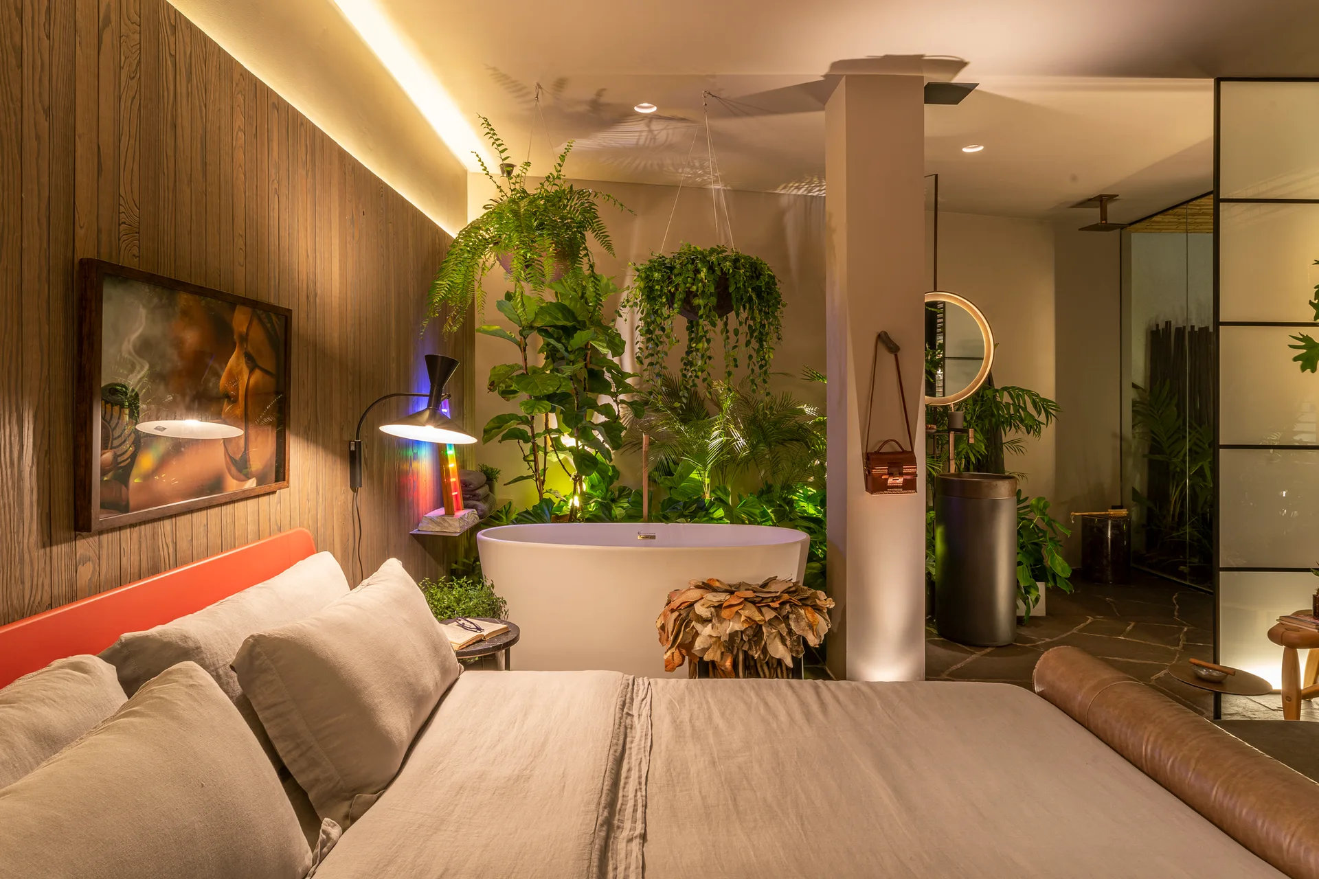 A contemporary bedroom features a grey linen bed, a white freestanding bathtub, and a pedestal sink surrounded by dense hanging and potted plants.