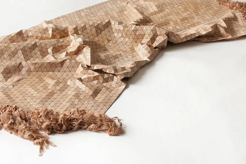 A tan textile sample with a structured three-dimensional geometric triangle pattern and loose frayed fringe borders lies on a white background.