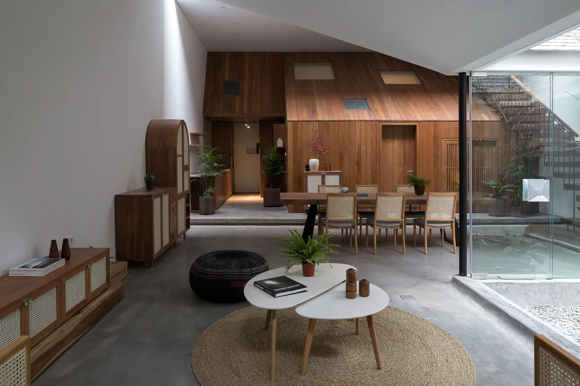 An open dining and living area features a wood-paneled sloped ceiling, dining table with cane chairs, concrete floors, and a glass partition.