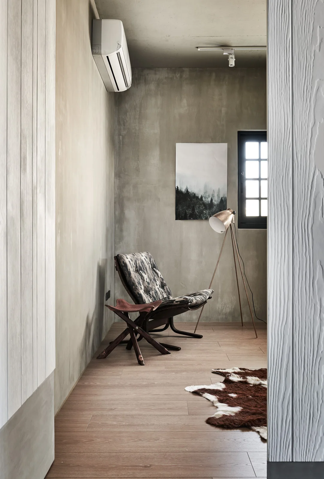 A minimalist reading nook features a camouflage-patterned lounge chair, a tripod floor lamp, and concrete-finish walls over wood flooring.