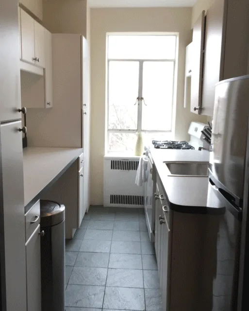 A narrow galley kitchen features off-white cabinetry, dark grey square floor tiles, stainless steel appliances, and a bright window at the far end.