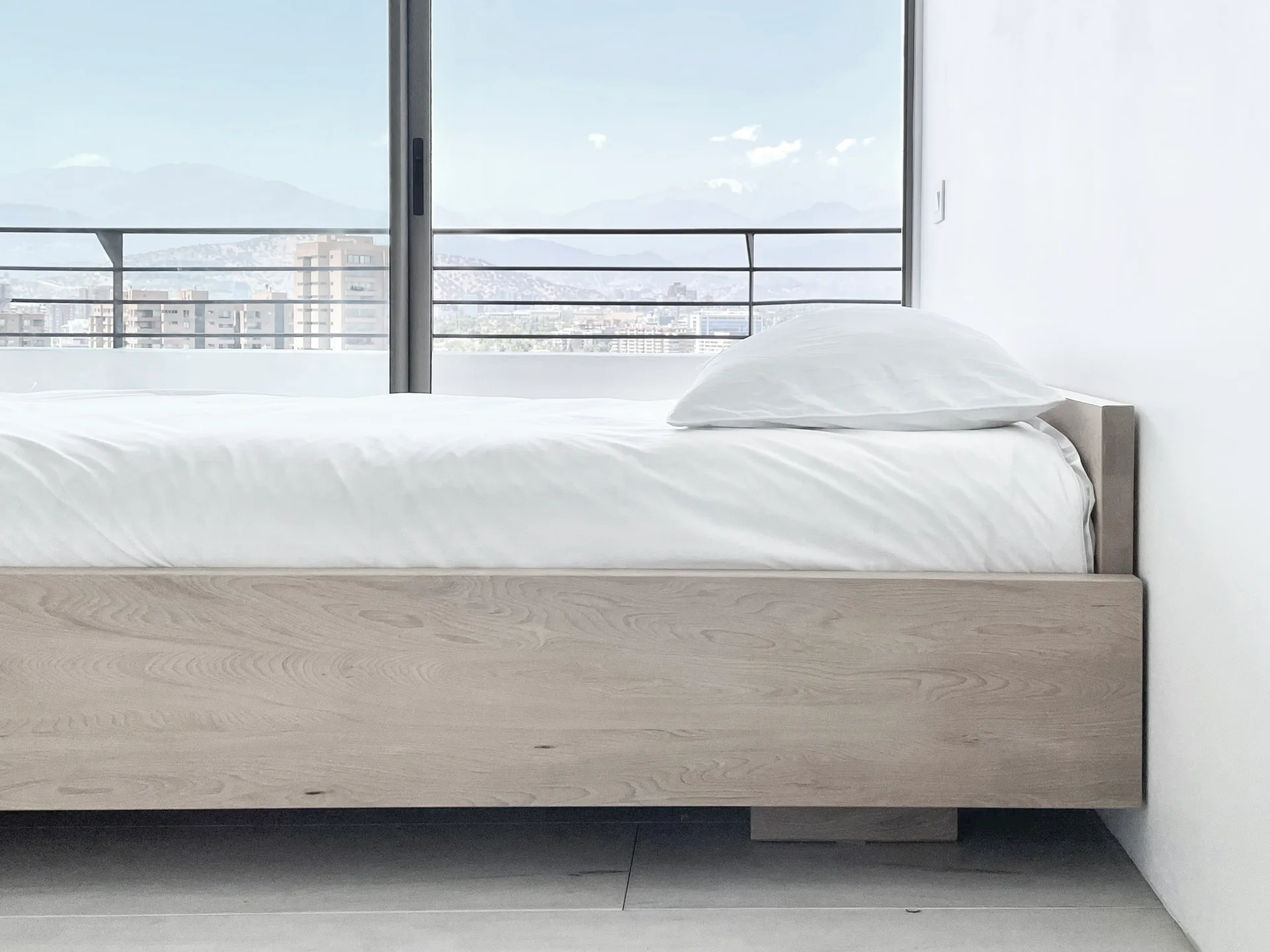 A minimalist bedroom featuring a light wood platform bed with white bedding next to a large window showing a city skyline.
