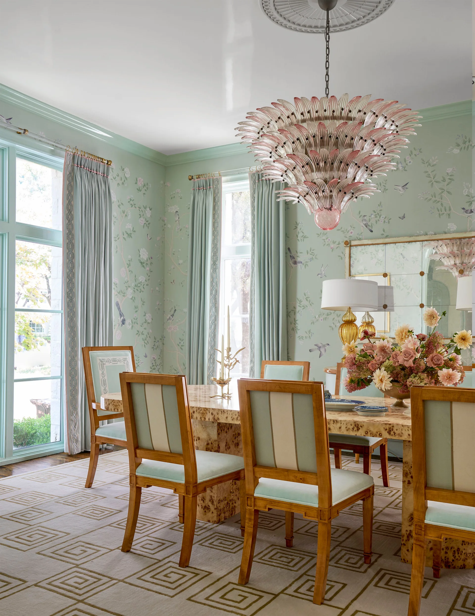 A formal dining room features a glossy burl wood table, mint green upholstered chairs, botanical wallpaper, and a large pink glass chandelier.