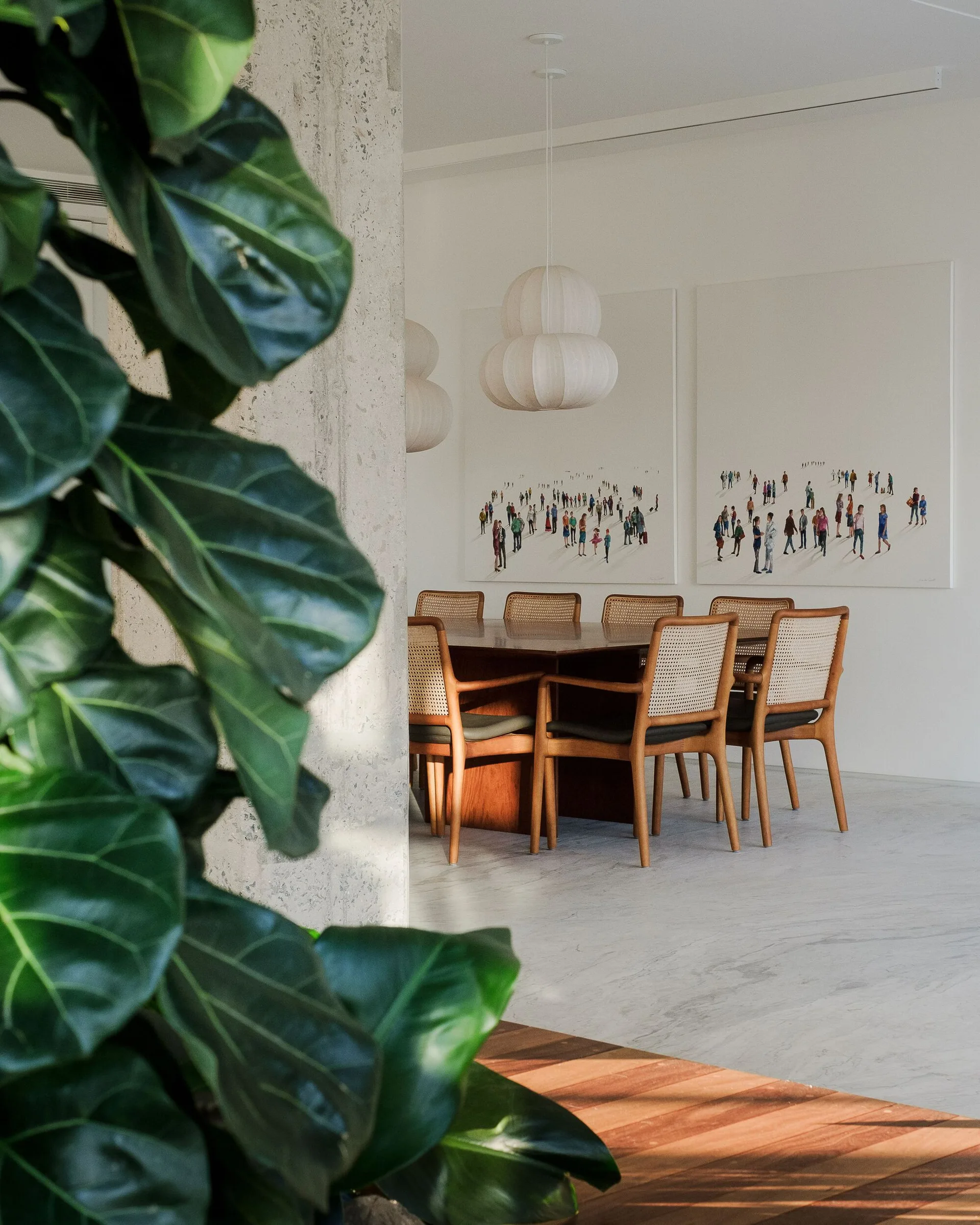 A modern dining room featuring a wooden table, cane-back chairs, and two cloud-like paper pendant lights beneath large paintings of people on white canvases.
