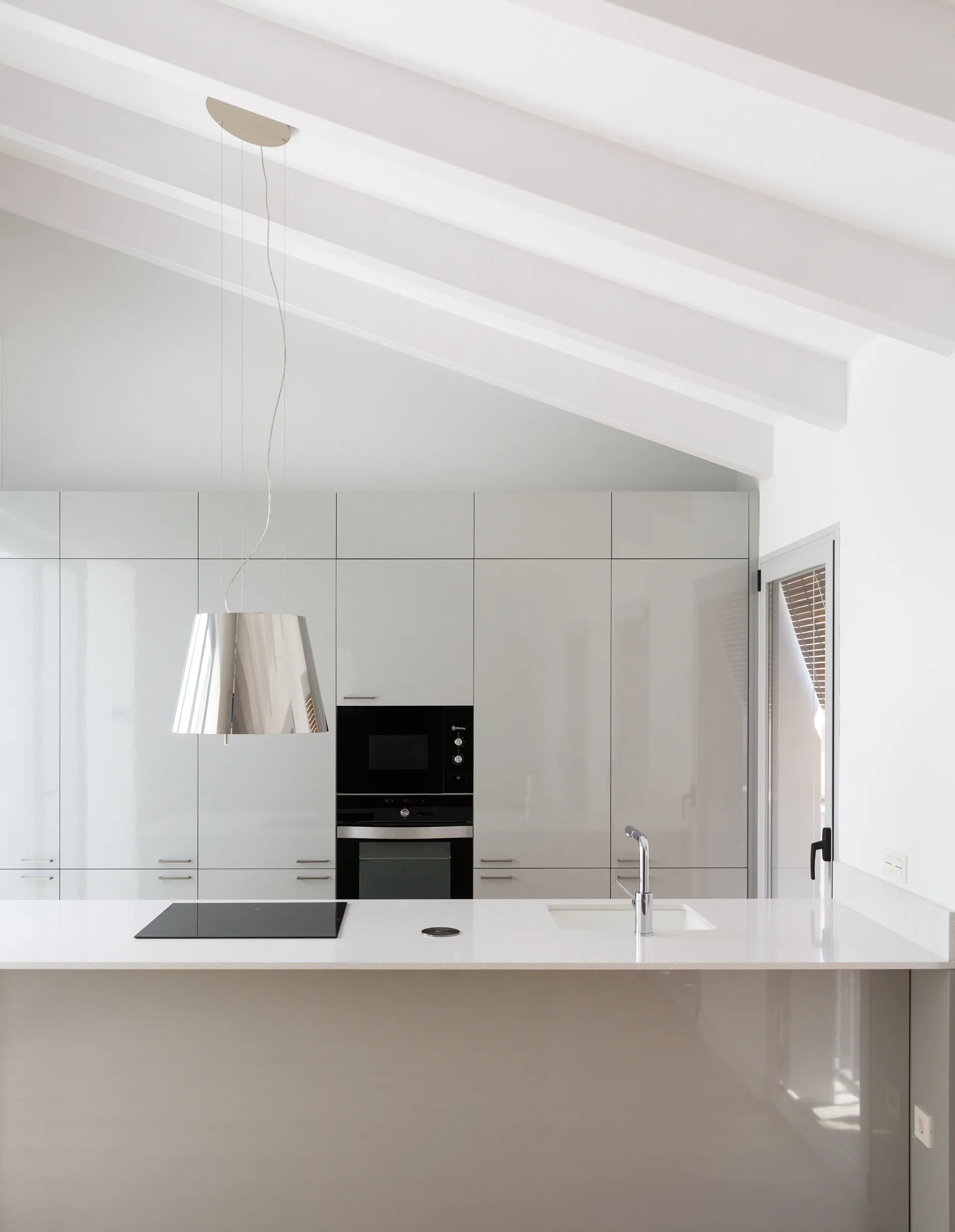 A minimalist kitchen features high-gloss taupe cabinets, a white island with a built-in cooktop, and an exposed white beamed ceiling.