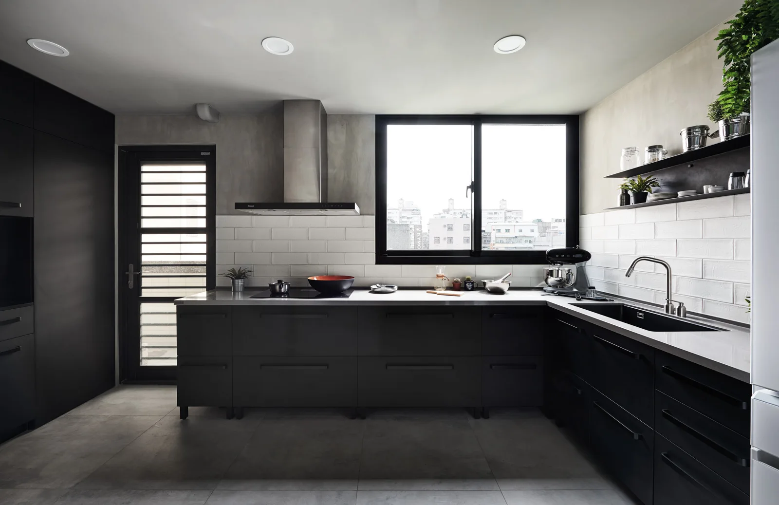 A modern kitchen with dark matte base cabinets, a white subway tile backsplash, concrete-like upper walls, and a large window above the grey countertop.