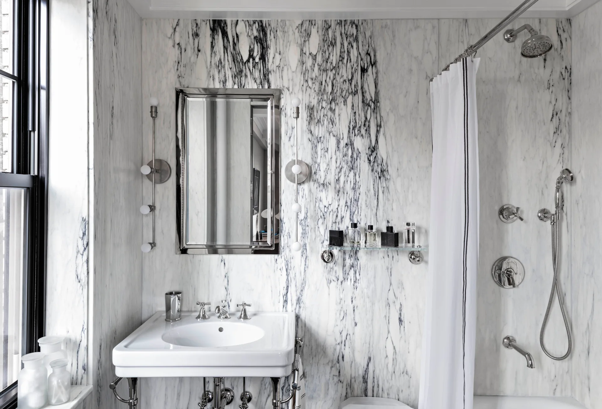 A white bathroom with black-and-grey veined marble walls, a chrome console vanity sink, vertical globe sconces, and a white shower curtain.