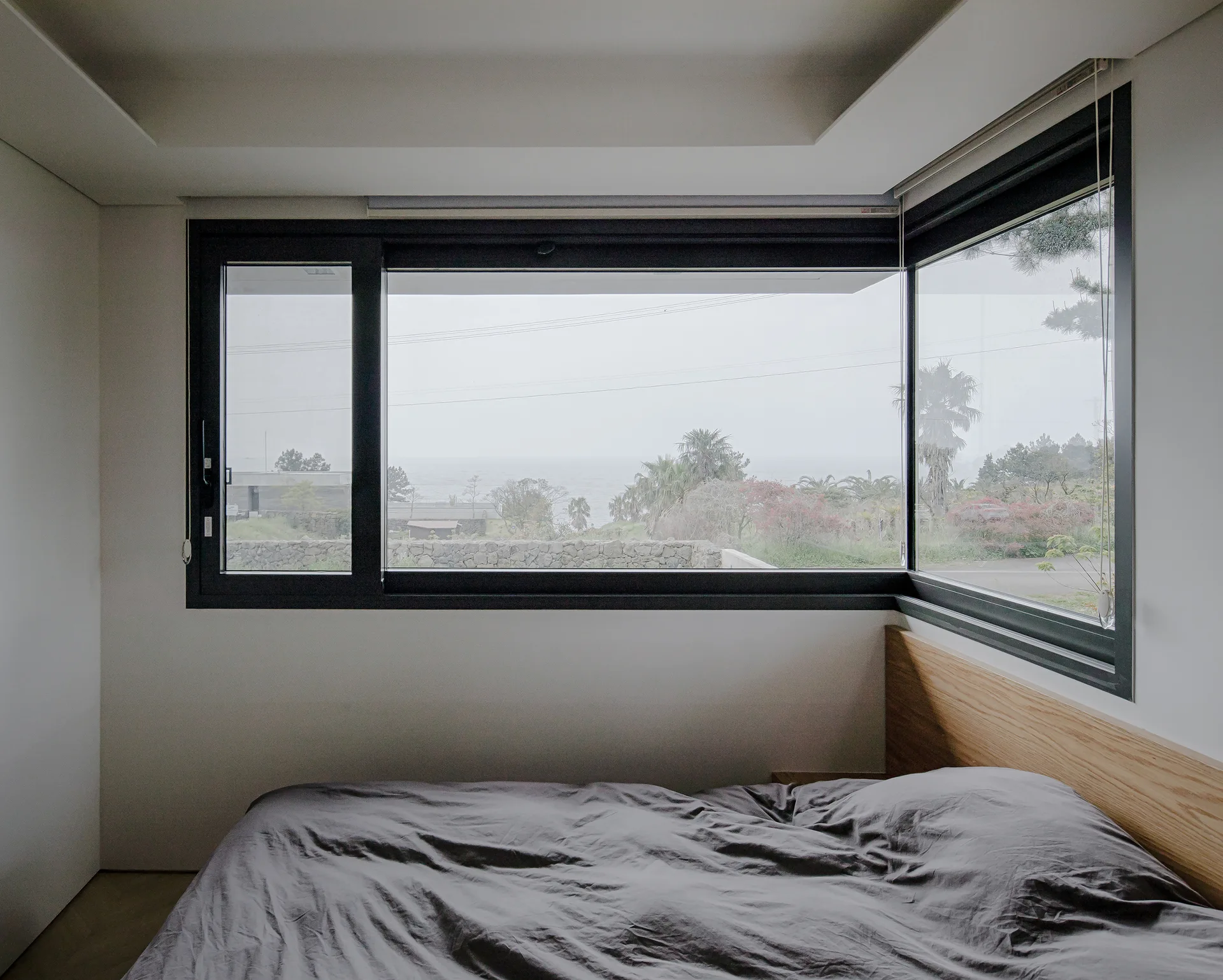 A minimalist bedroom with a low-profile bed, gray linens, a light wood headboard, and a large black-framed corner window showing an outdoor coastal view.