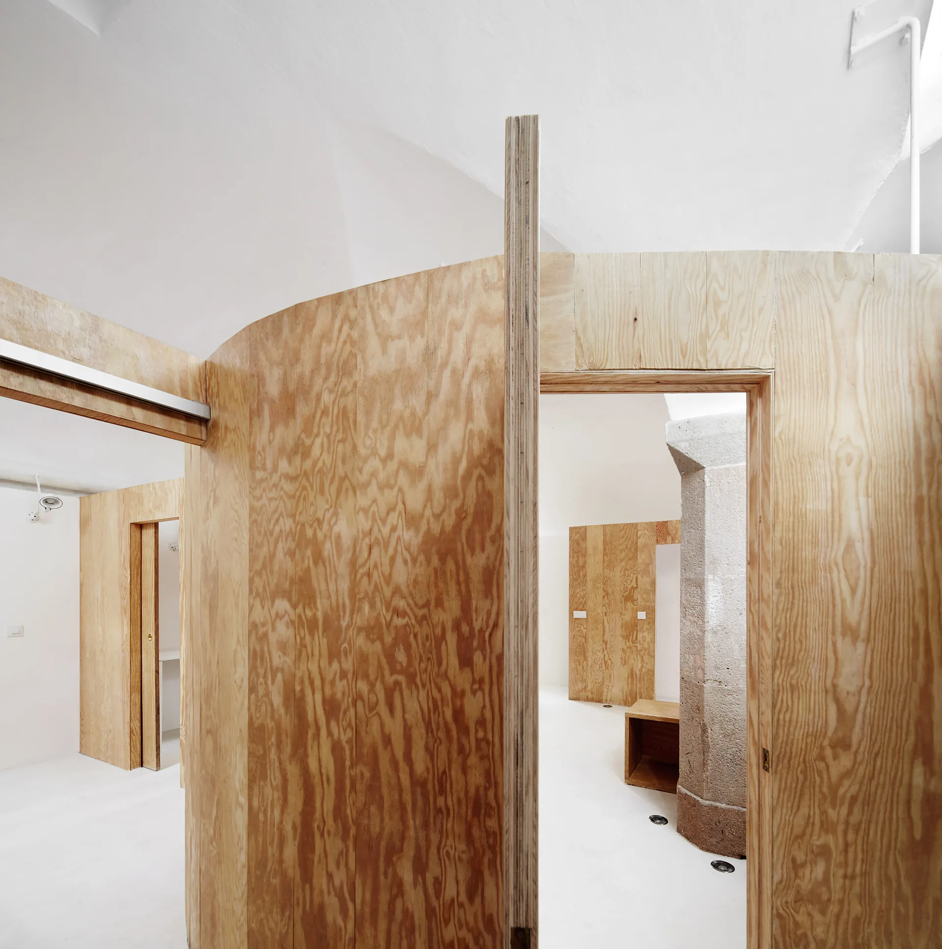Curving plywood walls with open doorways partition a minimalist room with white floors and an exposed stone pillar.