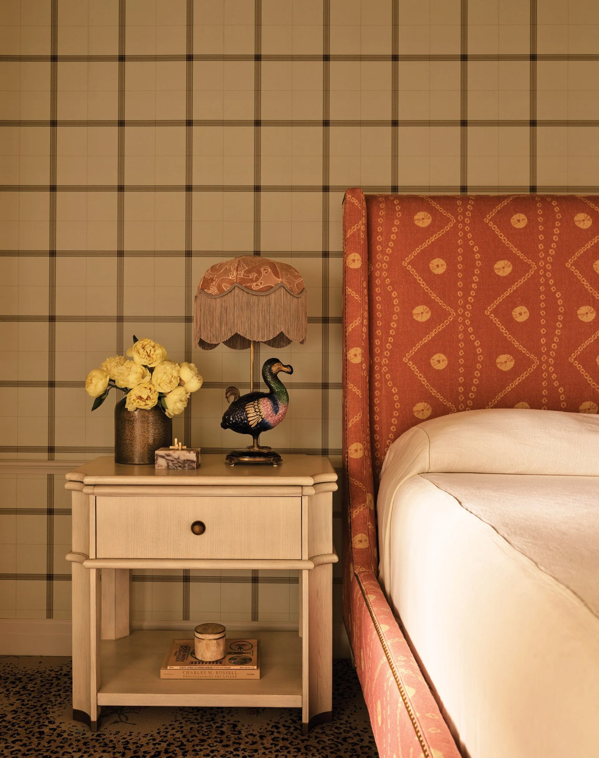 A light wood nightstand holds a whimsical bird lamp next to an orange patterned headboard against plaid wallpaper in an eclectic bedroom.