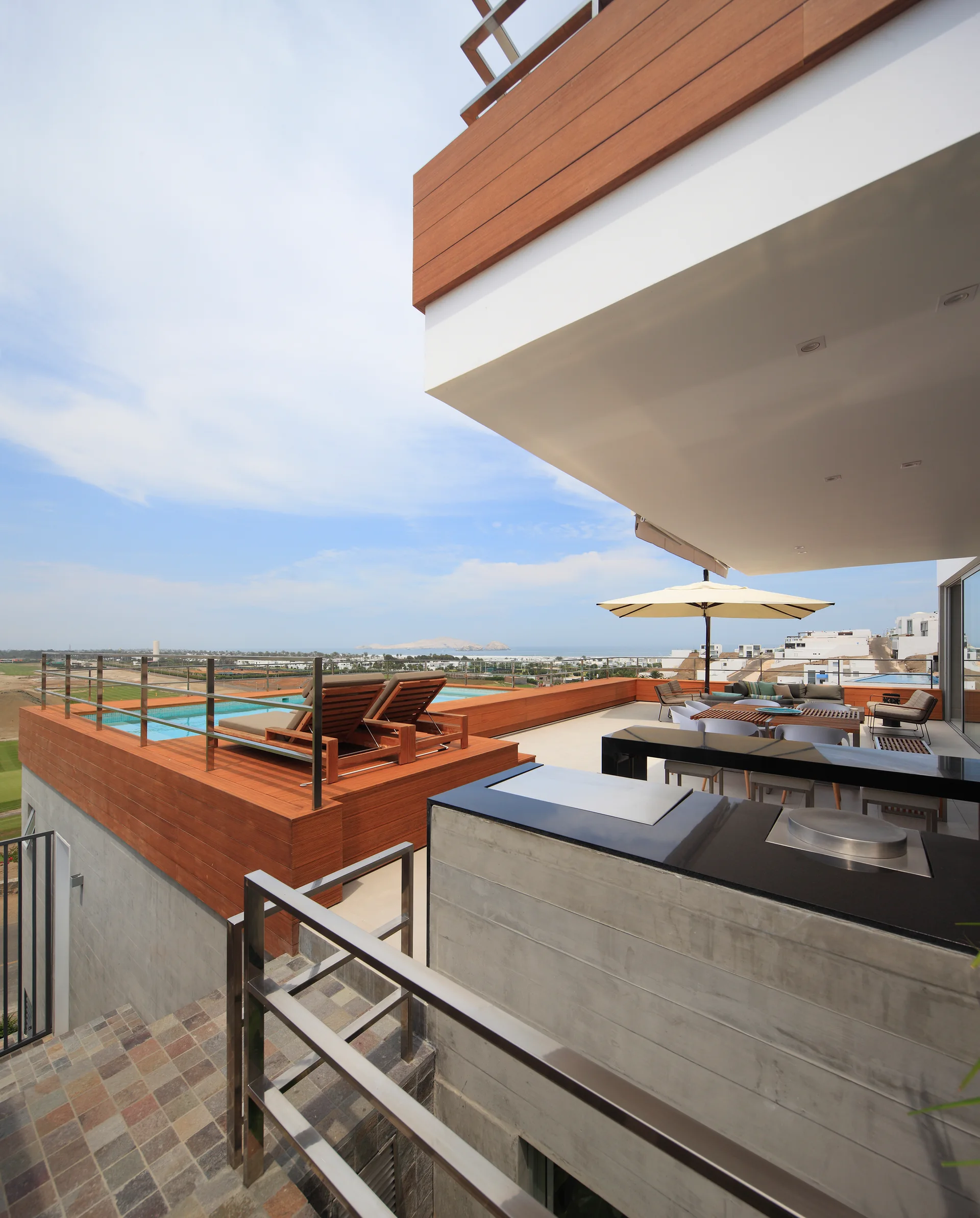 An elevated concrete terrace features a wood-clad swimming pool with lounge chairs, an outdoor kitchen, and a dining area under a large umbrella.