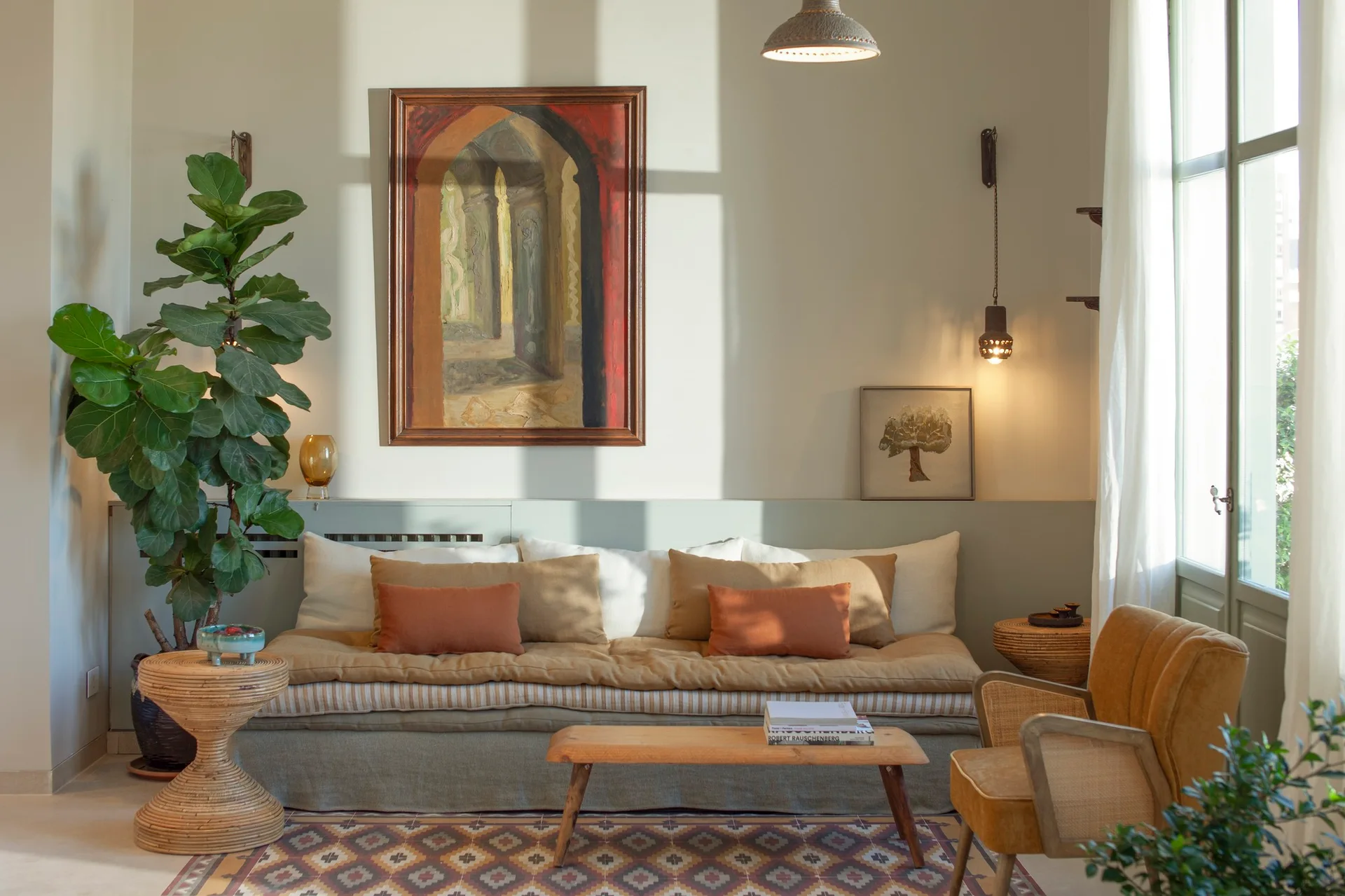 A sunlit living room featuring a layered sofa with terracotta pillows, a low wooden coffee table, an armchair, and a large framed painting.