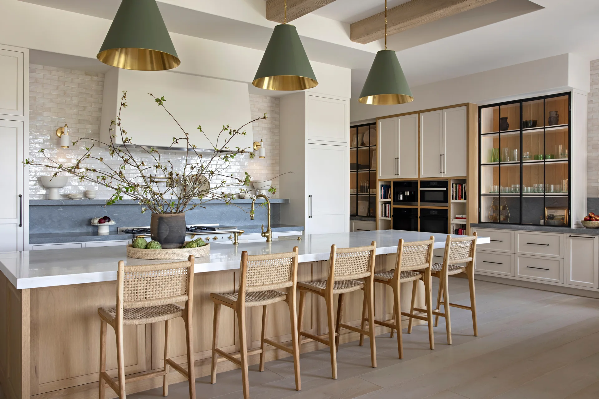 A spacious kitchen features a light oak island with woven bar stools, topped by three green conical pendant lights and a white stone countertop.