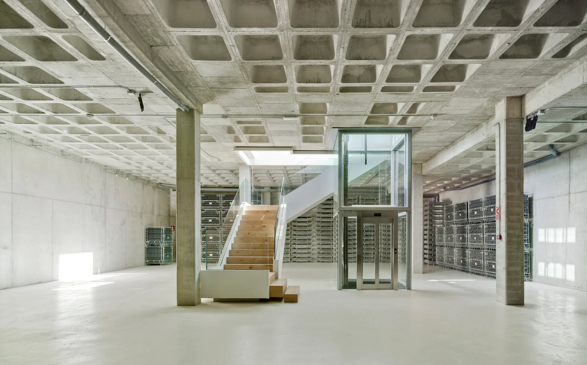 A large concrete interior featuring a prominent waffle slab ceiling, a central wooden staircase with glass railings, and a glass elevator.