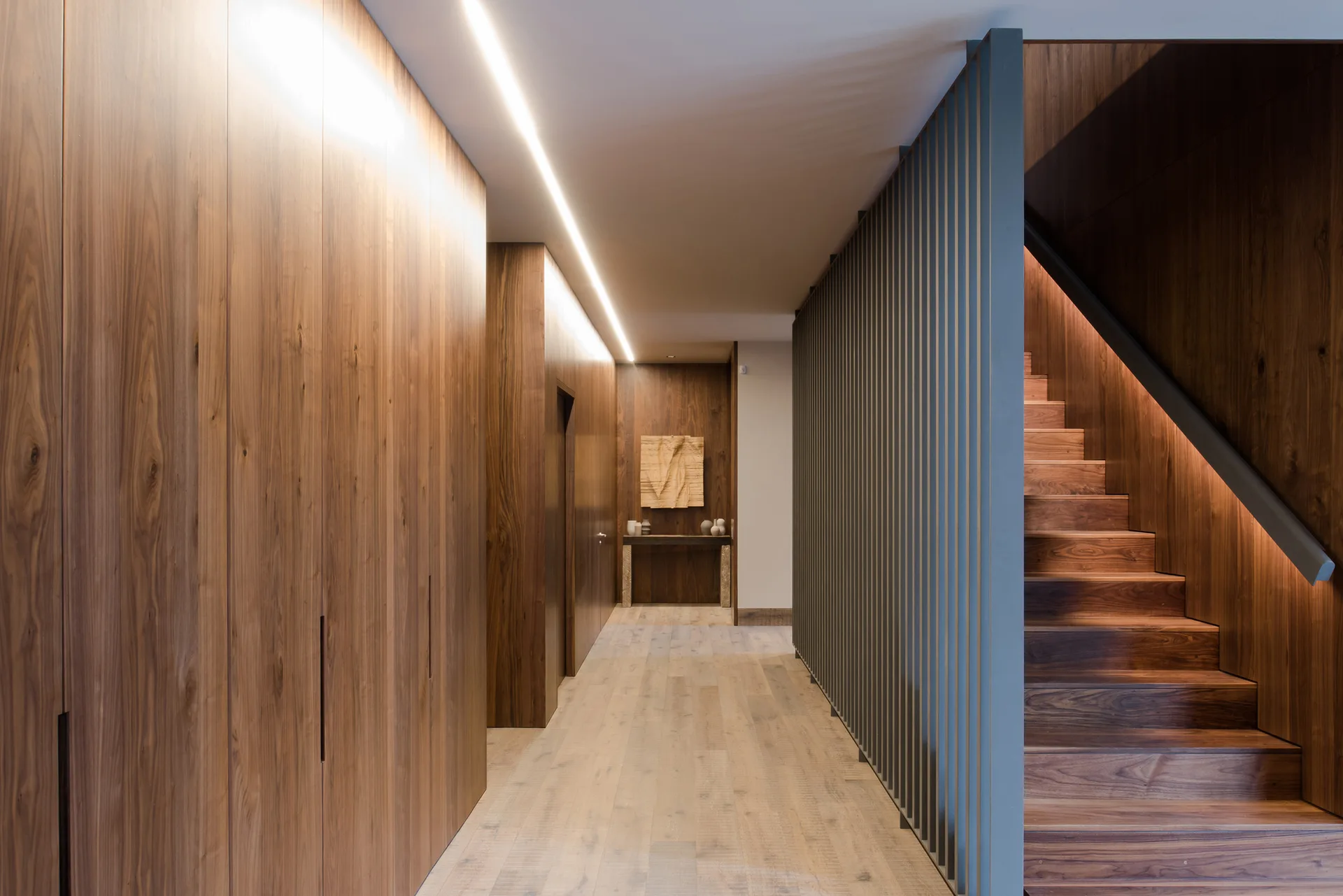 A contemporary hallway features floor-to-ceiling wood paneling, a wooden staircase behind a dark slatted screen, and a recessed ceiling light strip.