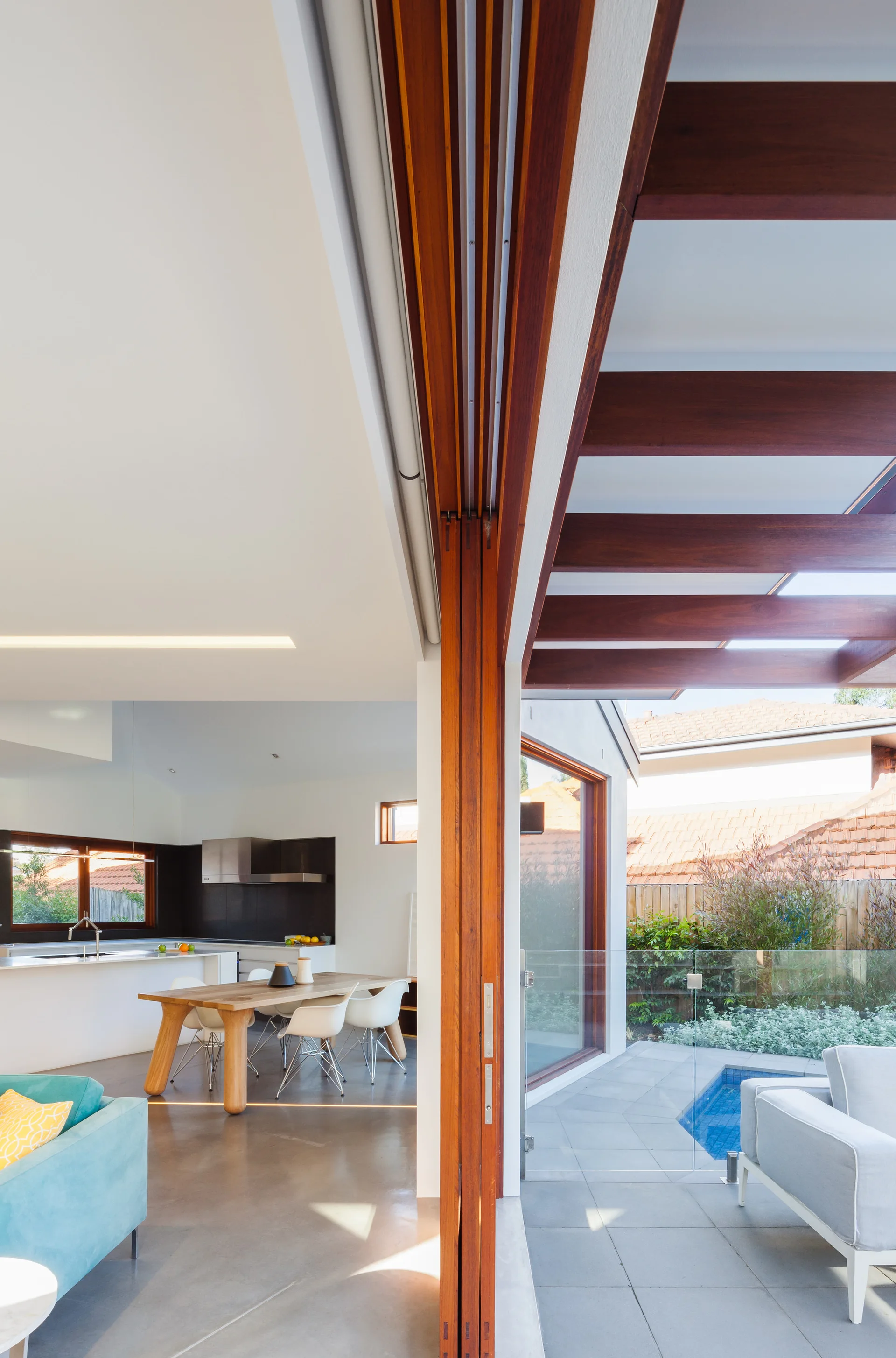 An open-plan dining room with a wooden table leads to an outdoor patio and small swimming pool through a large wooden sliding door.