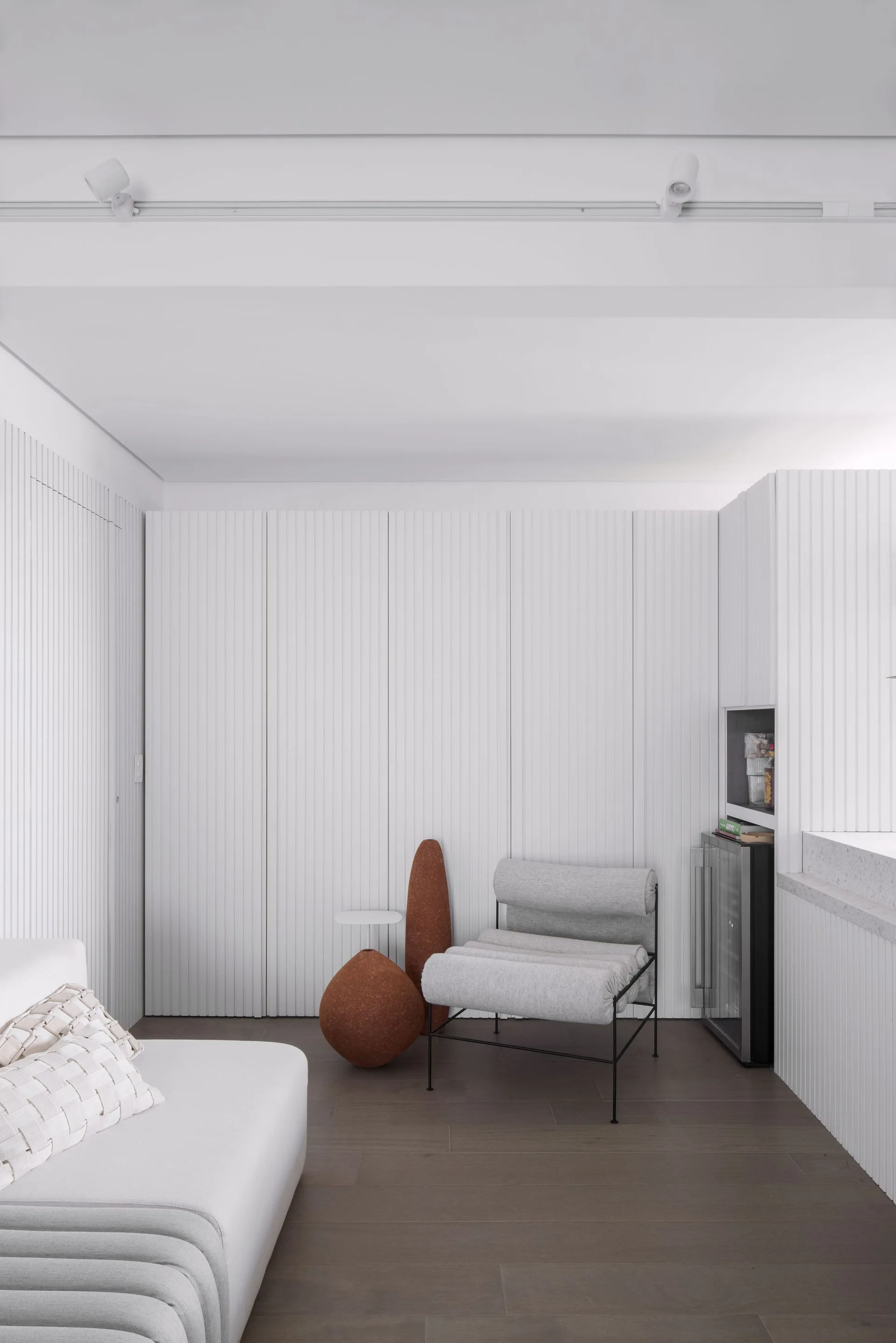 A minimalist room featuring white vertical paneled walls, a grey lounge chair, and two organic terracotta sculptures.