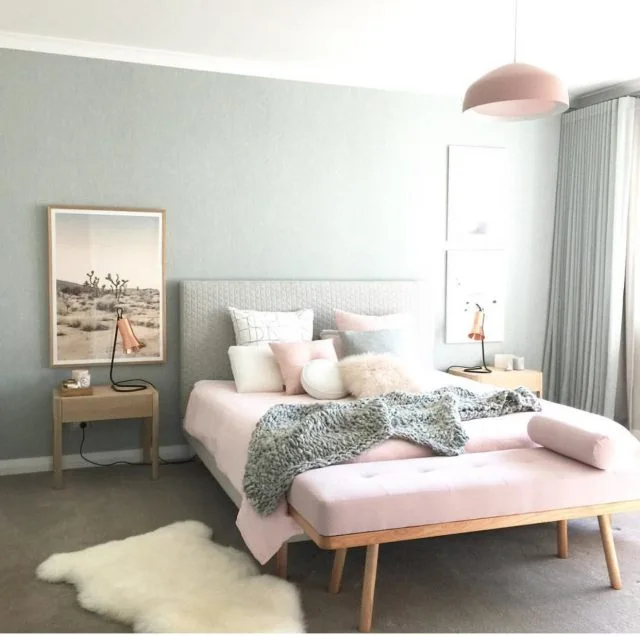 A bedroom features an upholstered bed with pink and grey bedding, light wood nightstands, a pink bench, and a muted blue-grey wall.