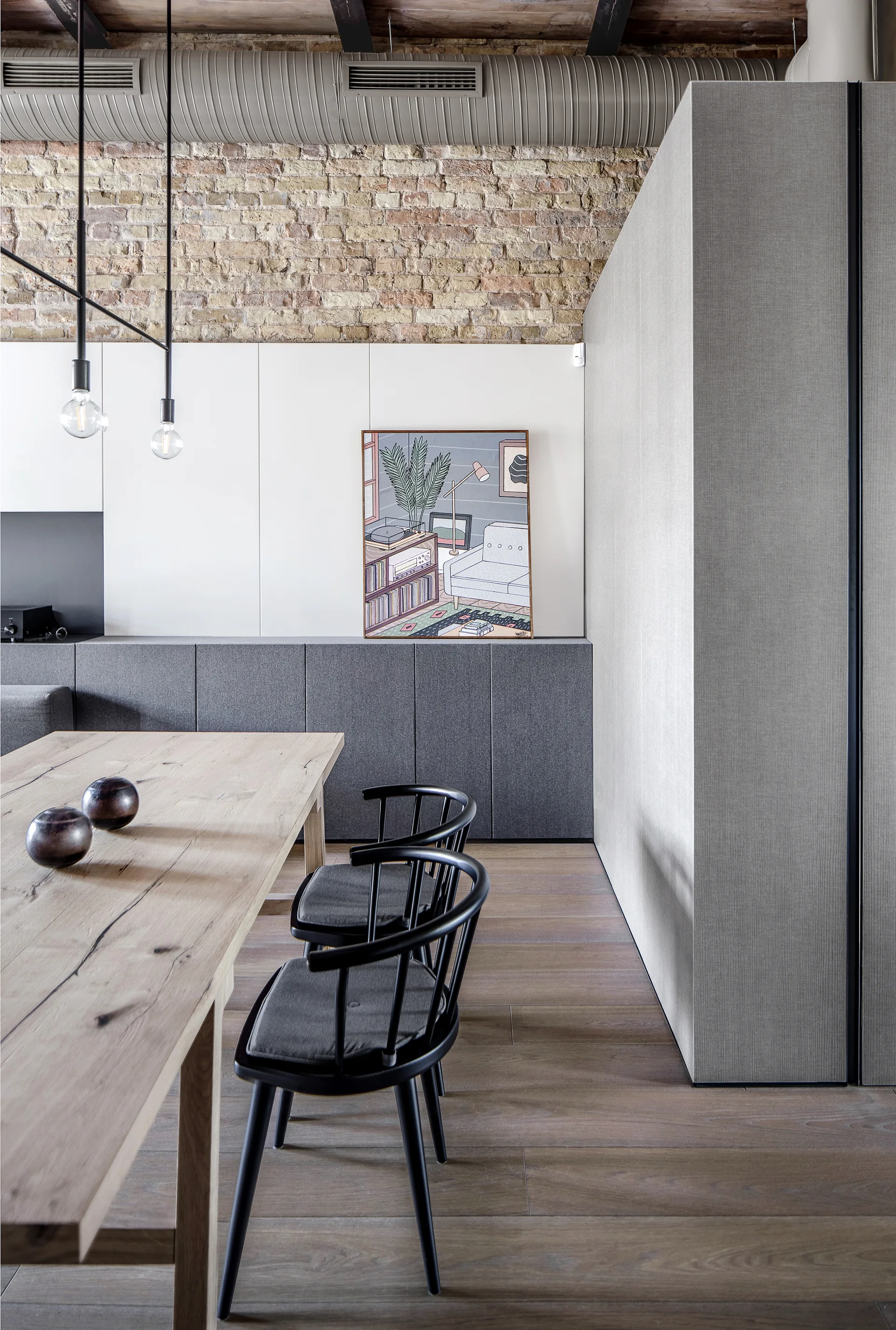 A light wood dining table with black spindle chairs stands before a wall of white paneling, grey cabinets, and upper exposed brick.