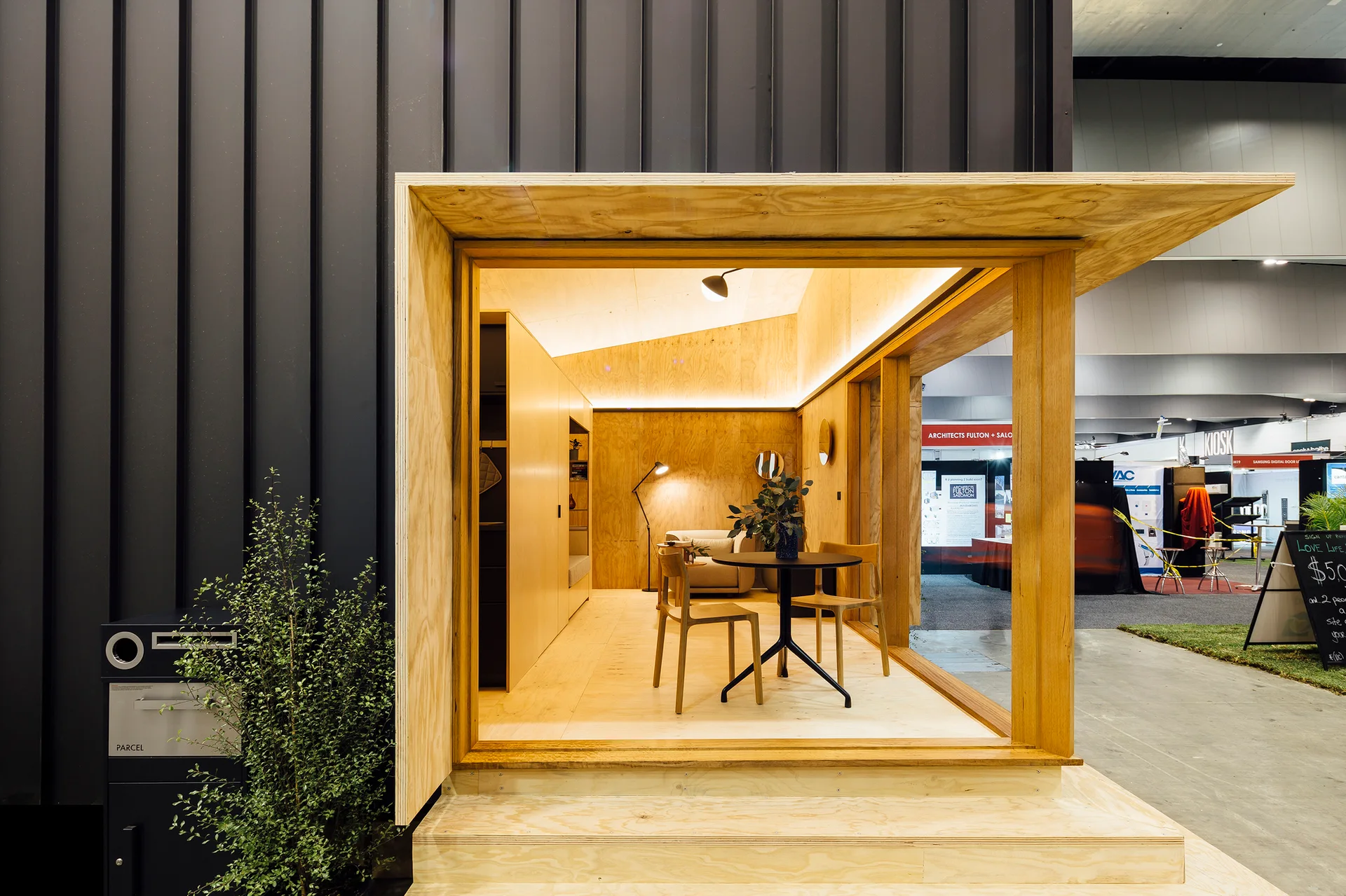 A modern modular pod features a light plywood interior with a black dining table, chairs, and a built-in cabinet against a dark exterior wall.