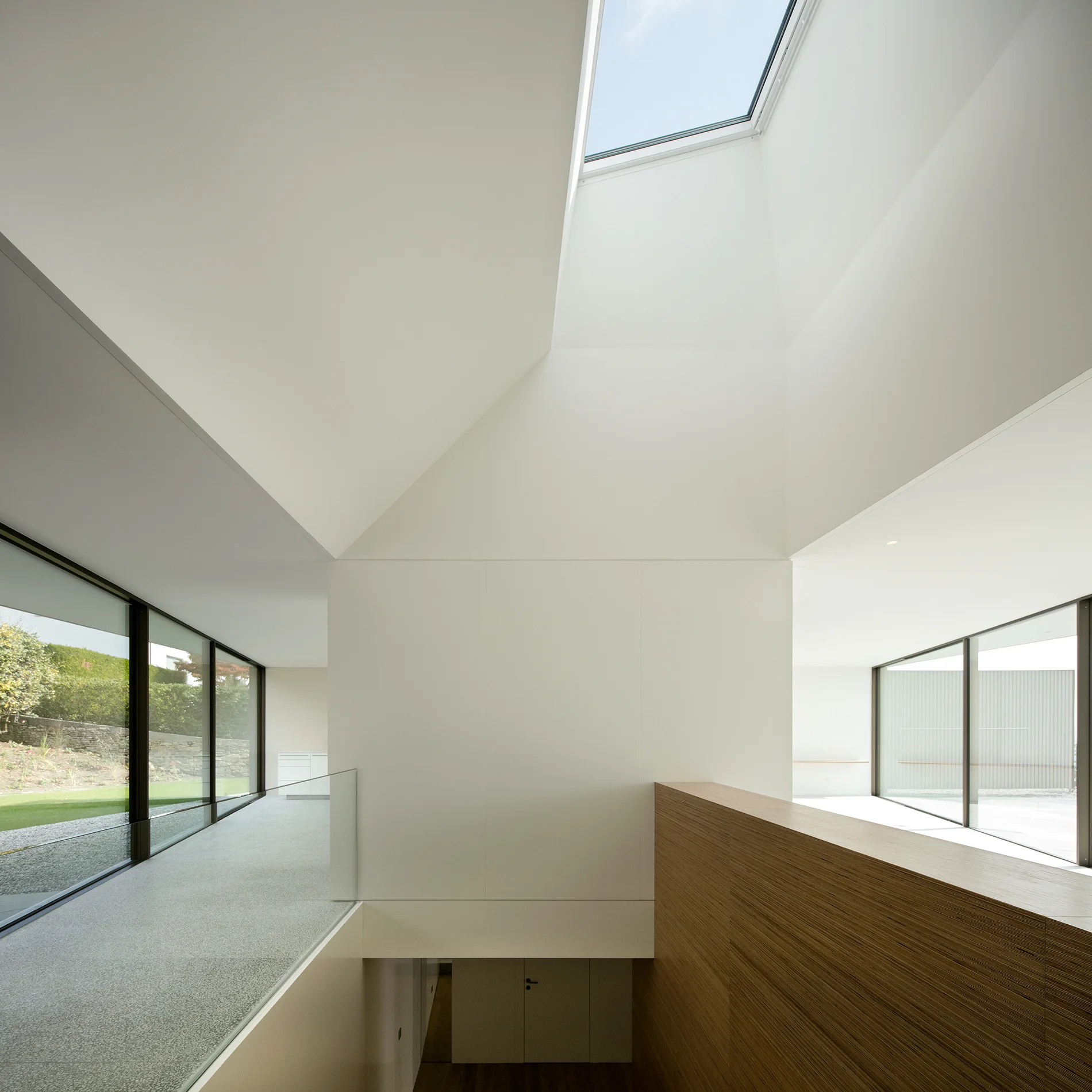A minimalist hallway features a large rectangular skylight, floor-to-ceiling glass windows, a glass railing, and horizontal wood wall paneling.