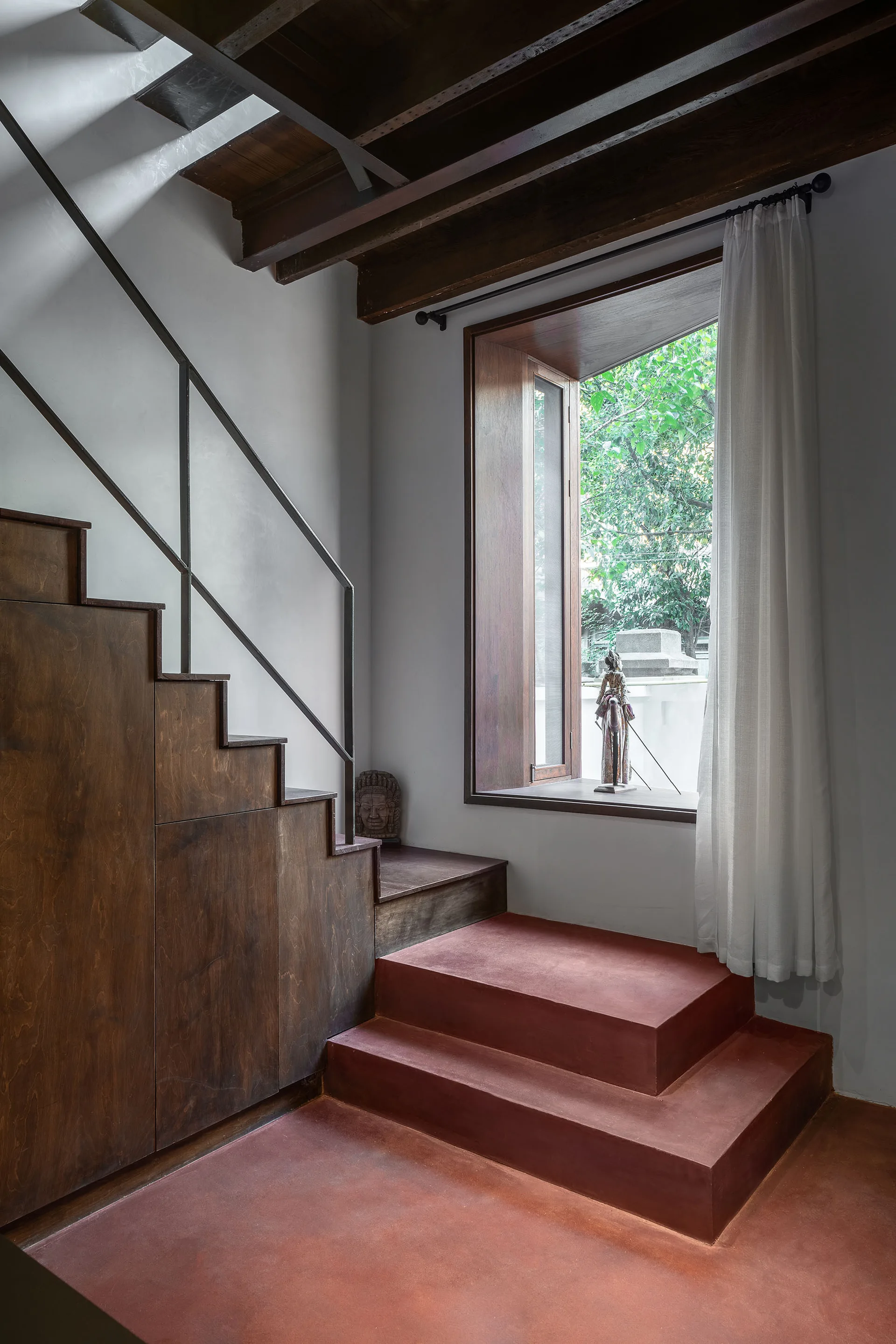A modern wood staircase with integrated storage cabinets meets red concrete steps next to a large window showing garden greenery.