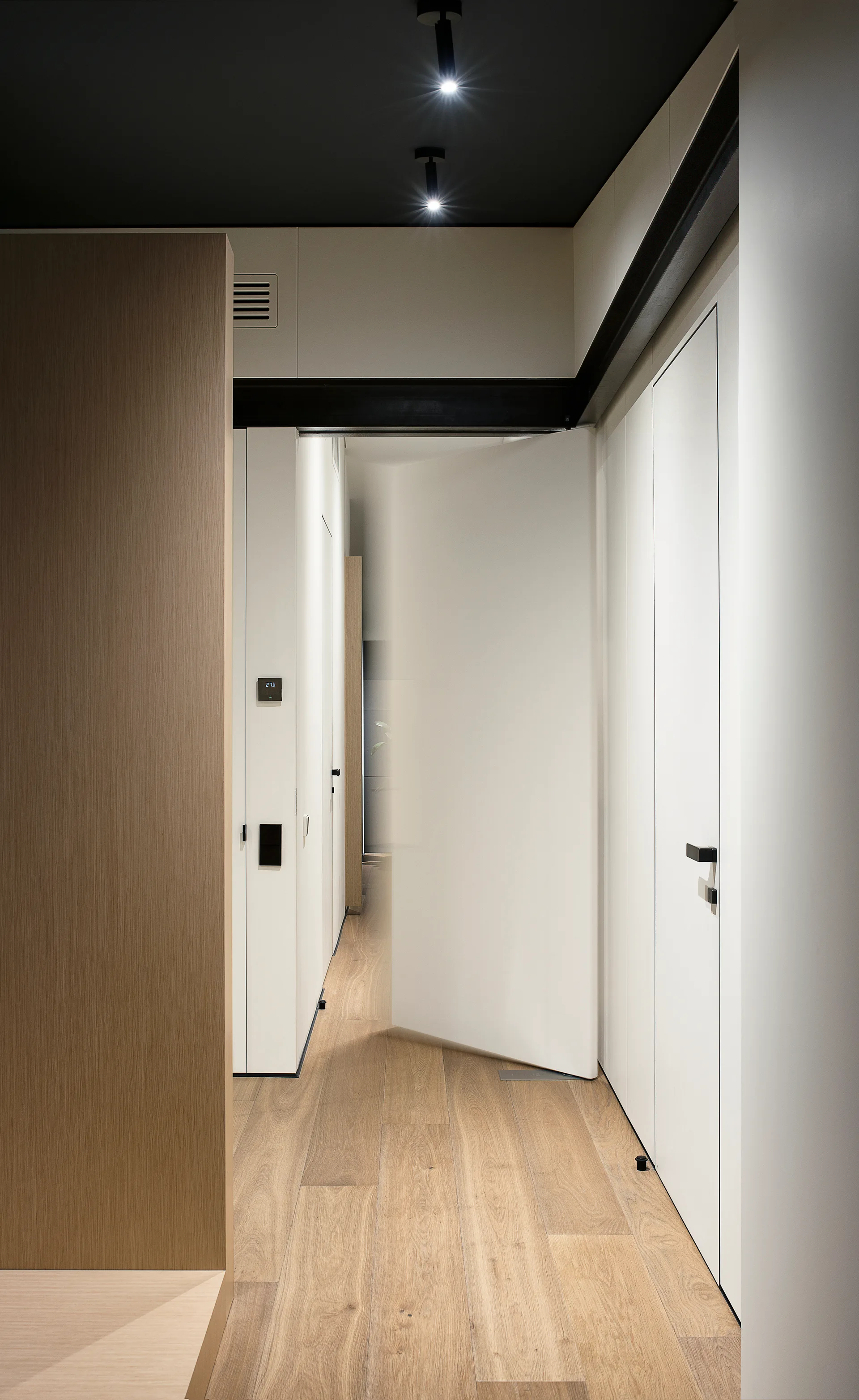 A modern minimalist hallway featuring light wood plank flooring, flush white doors with black handles, and a contrasting black ceiling with spotlights.