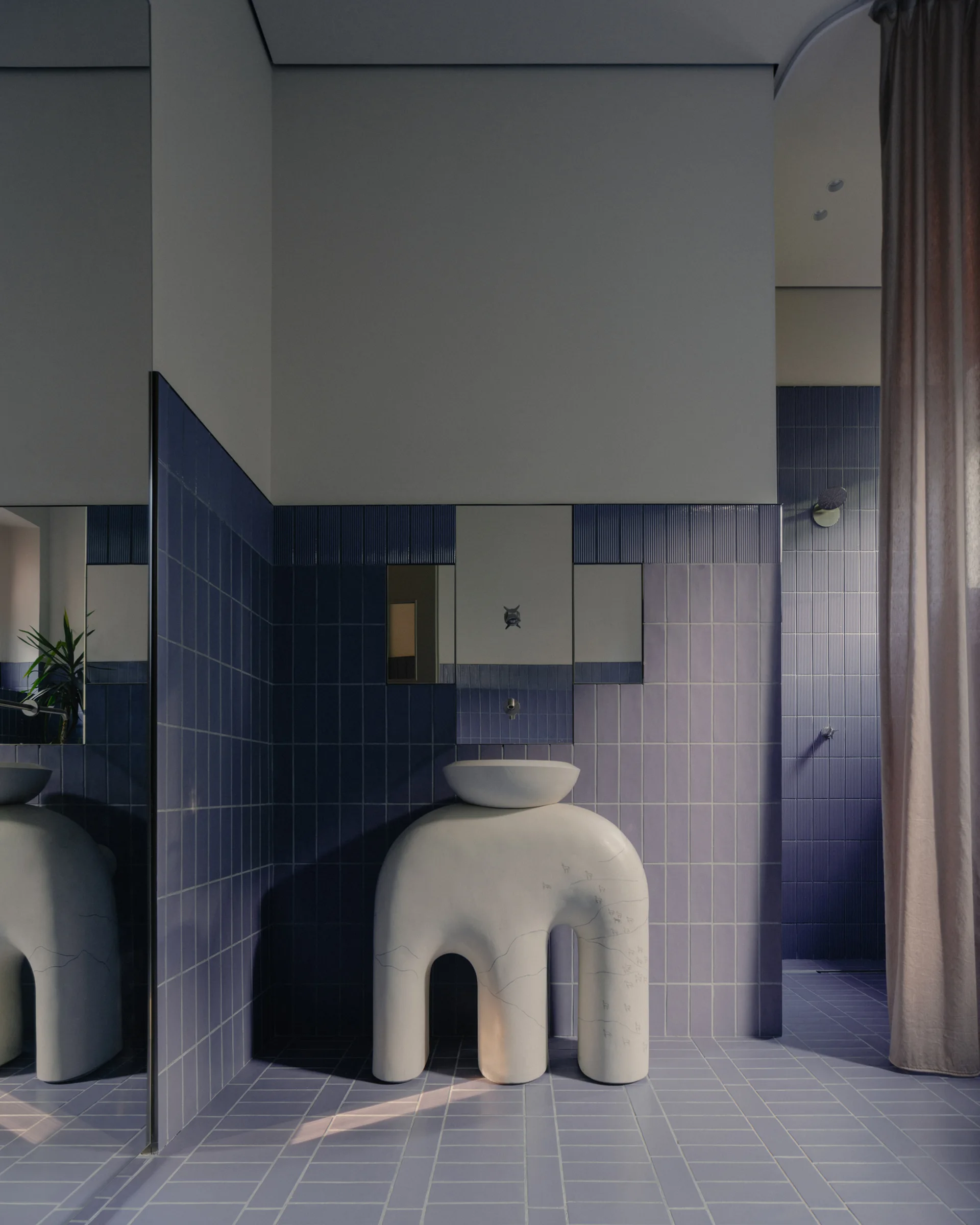 A minimalist bathroom features light blue tiled walls and floor, a white sculptural arch washbasin, and a tall mirror reflecting the tiled space.