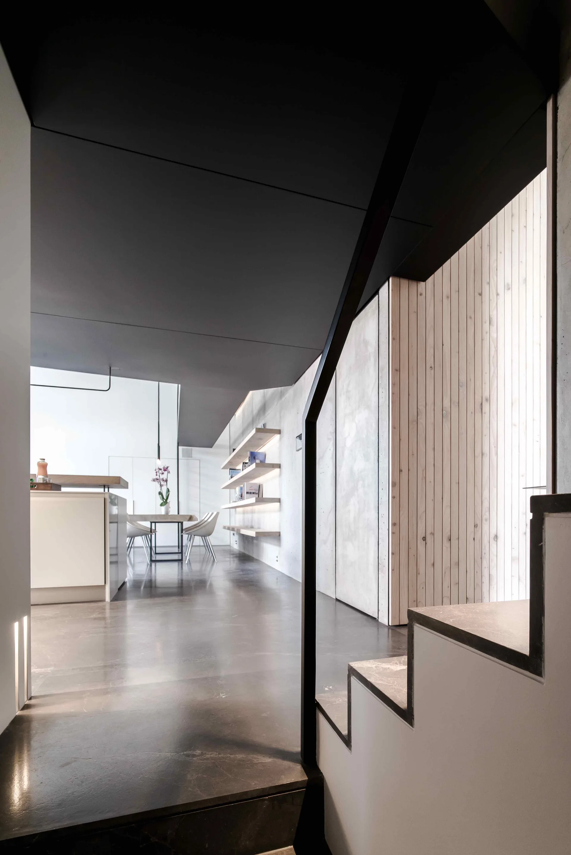 A view from a minimalist staircase showing a black ceiling, concrete walls with illuminated wood floating shelves, and a dining area in the background.