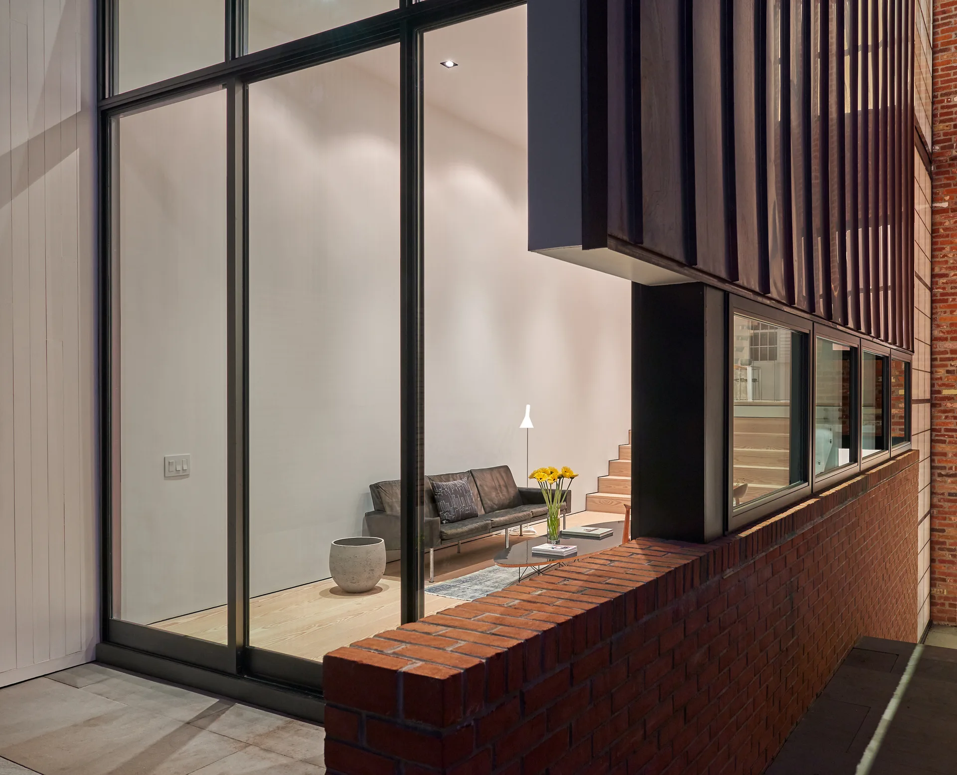 A modern living room with a black leather sofa and light wood floors is viewed through a large multi-pane glass window bordered by a brick wall.