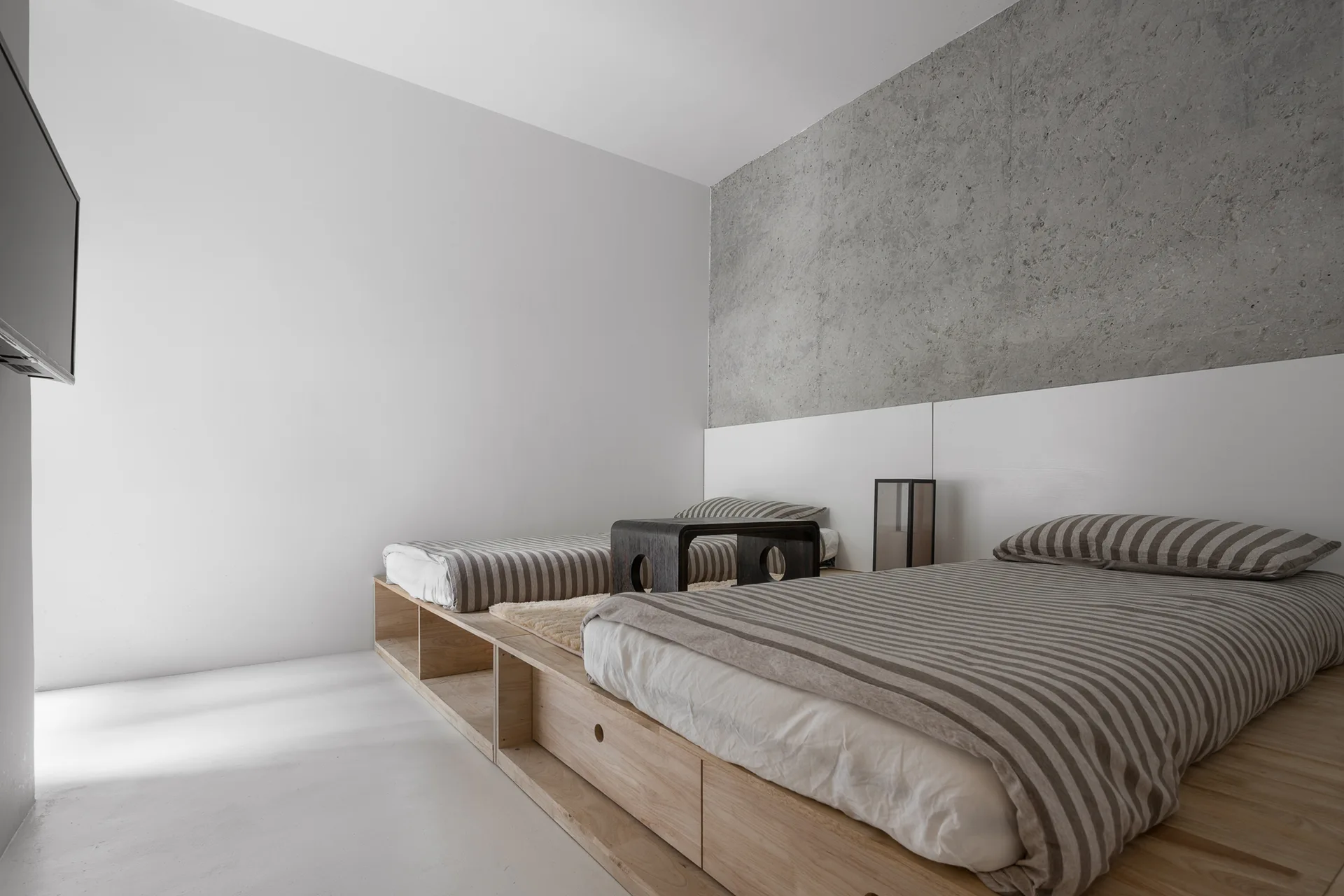 Two twin beds with striped linens are built into a low light wood platform base against a gray concrete accent wall.