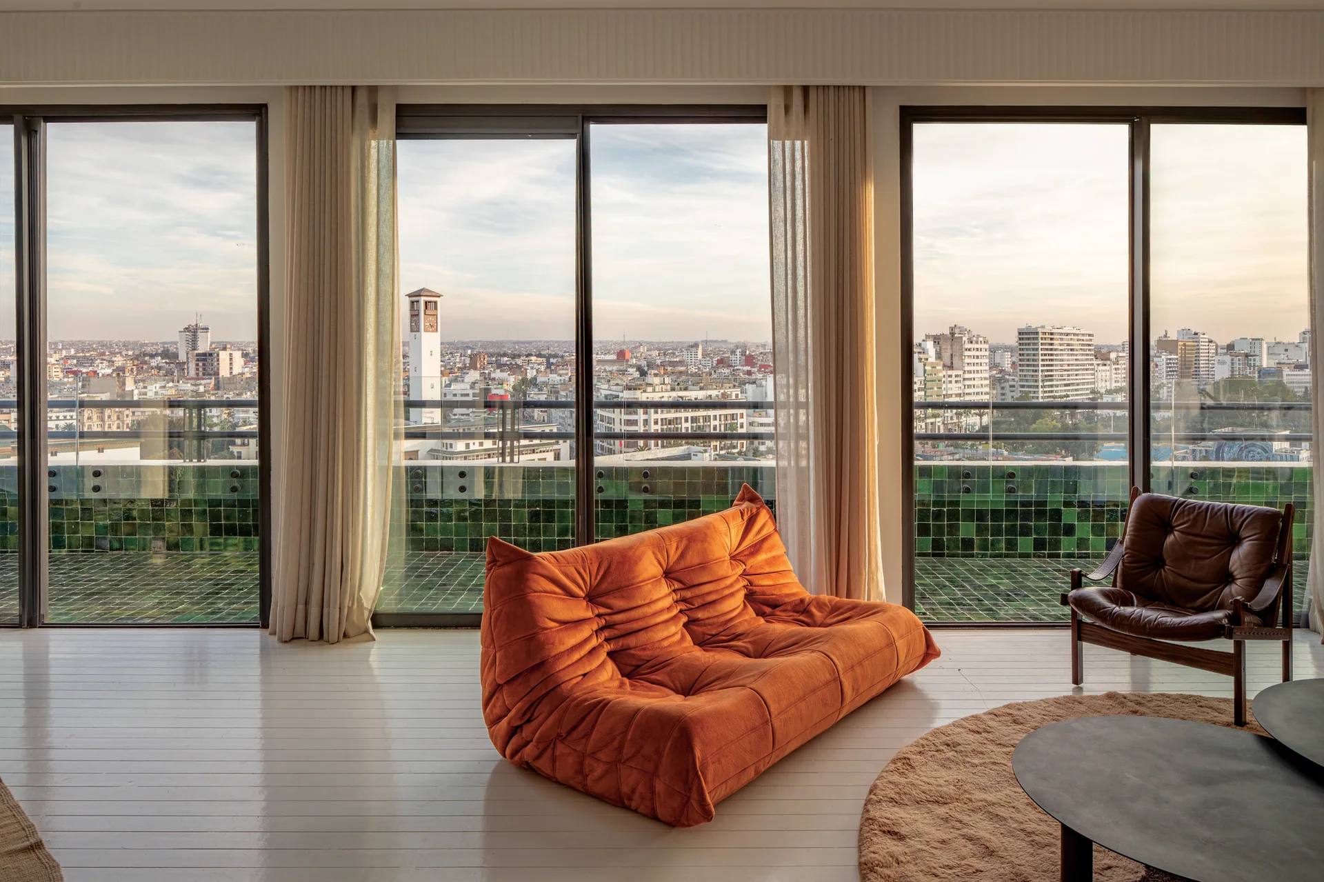 A bright living room features a low-profile orange tufted sofa and a brown leather chair facing large glass windows with city views.
