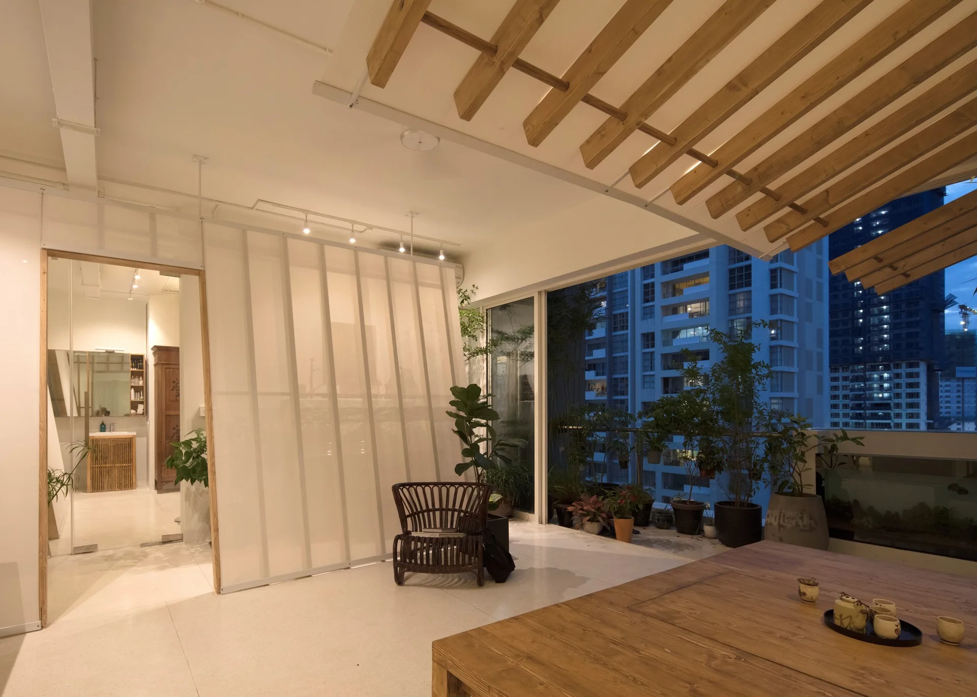 A covered apartment terrace at dusk featuring a large wood dining table, a slatted timber ceiling, and potted plants overlooking a city skyline.