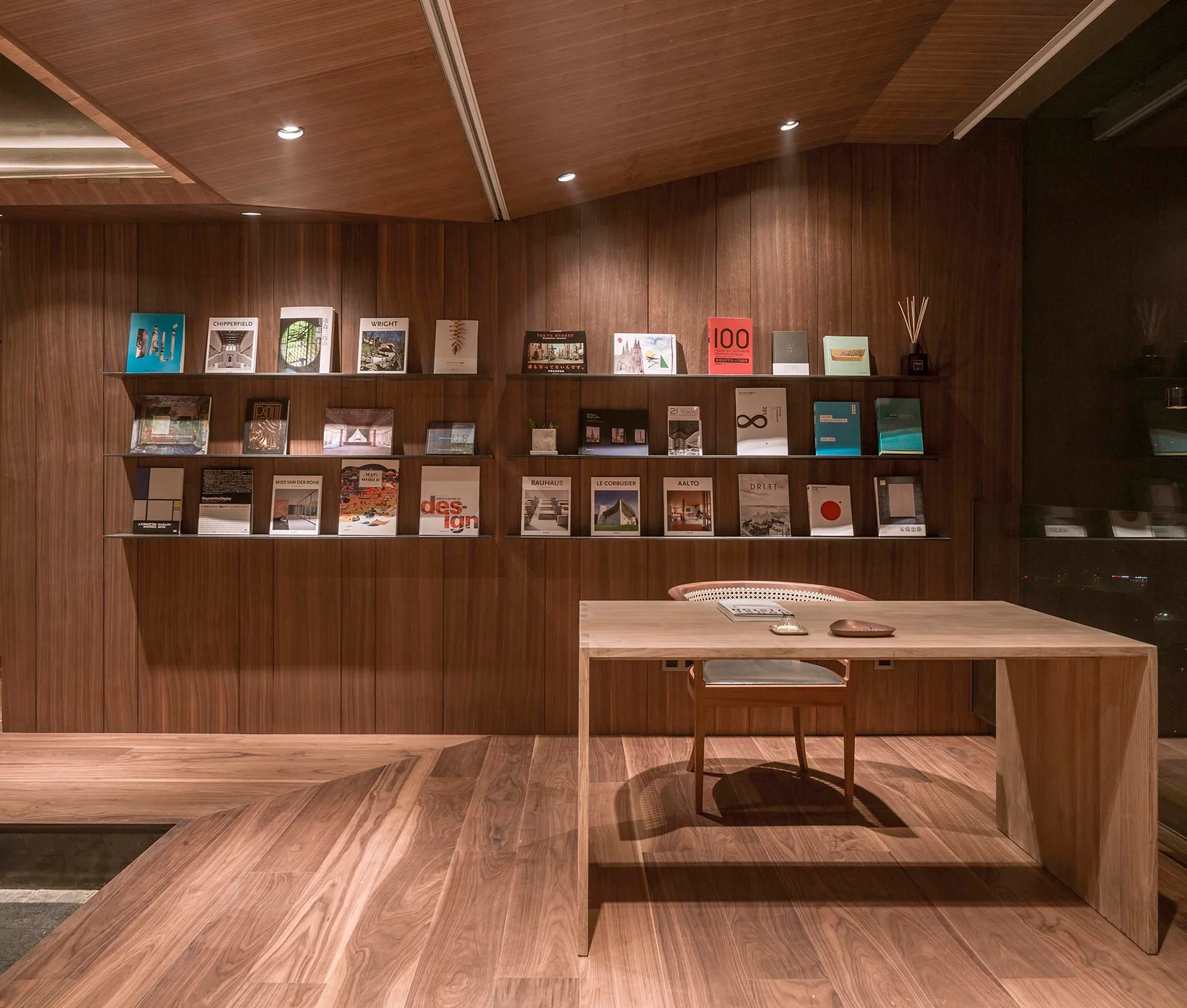 A minimalist study features wood-paneled walls and ceiling, a wooden desk and chair, and thin black ledge shelves displaying books face-out.