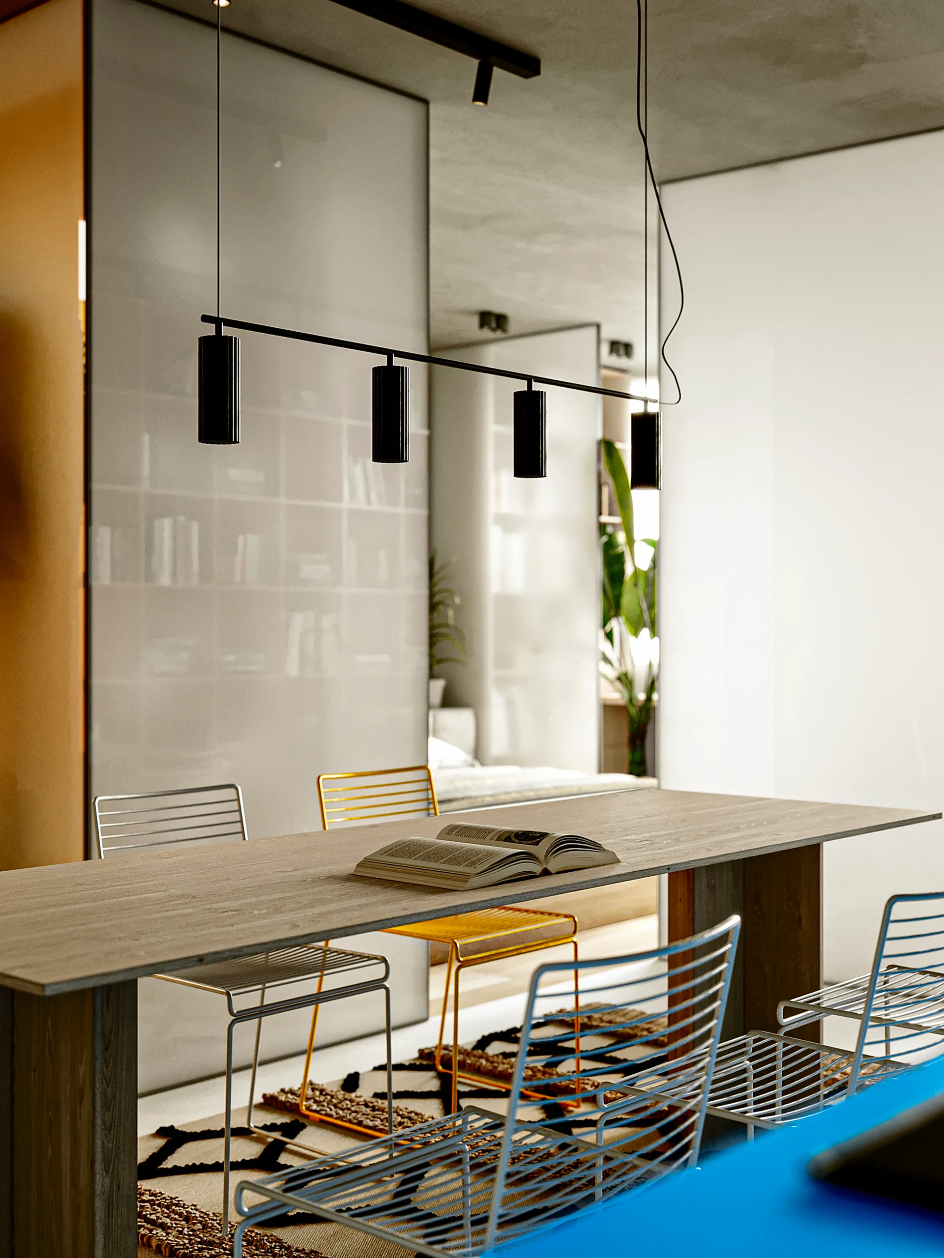 A long wooden dining table features colorful wireframe chairs and a black linear pendant light in a modern room with concrete ceilings.