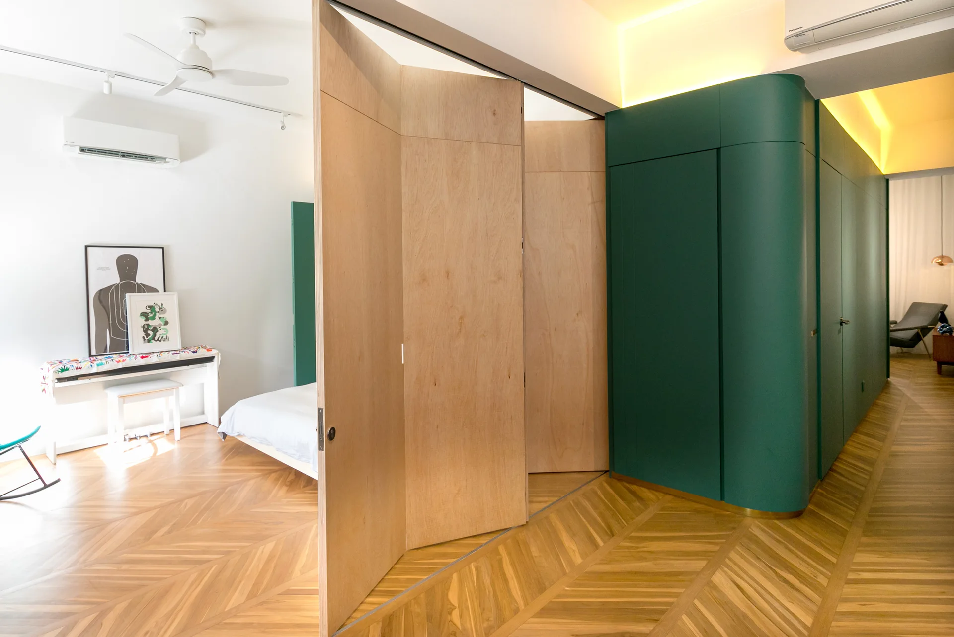 A modern bedroom features a folding wood partition wall, chevron wood flooring, and curved dark green built-in cabinetry with cove lighting.