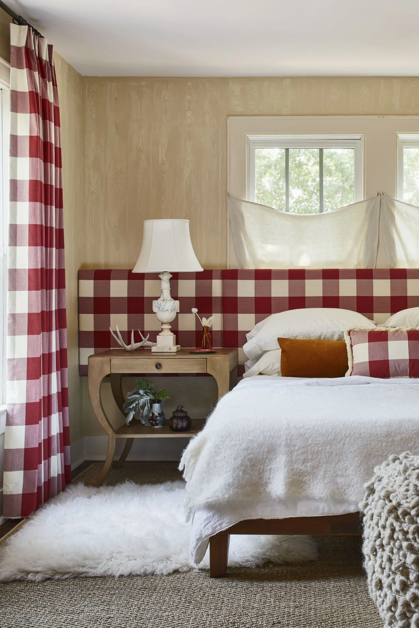 A bedroom features a bed with a red and white checkered headboard, a wooden nightstand with a lamp, and matching buffalo check curtains.